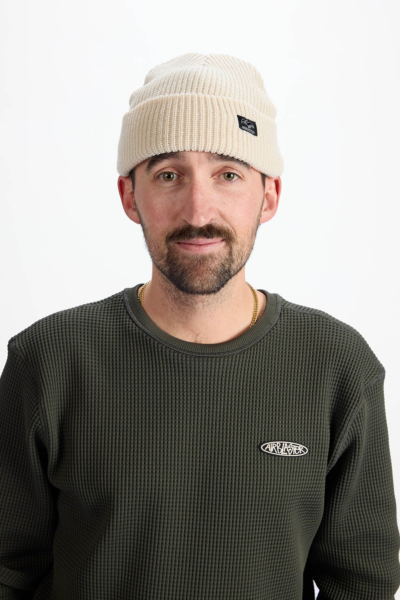 A man wearing a cream ribbed knit beanie with a small black logo patch and an olive green waffle-knit crewneck sweatshirt with a white chest logo, photographed against a plain white background.