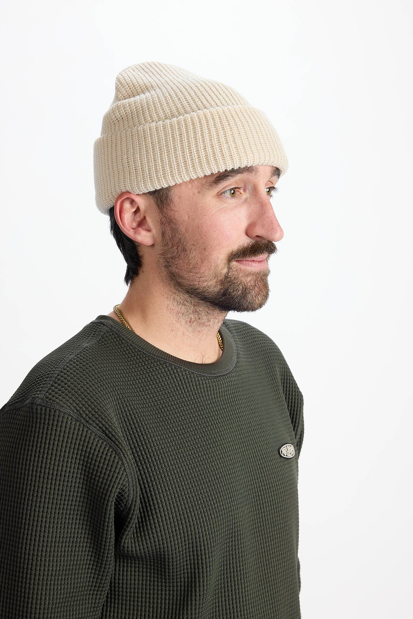 A man in profile wearing a cream ribbed knit beanie and an olive green waffle-knit crewneck sweatshirt, photographed against a clean white studio background.