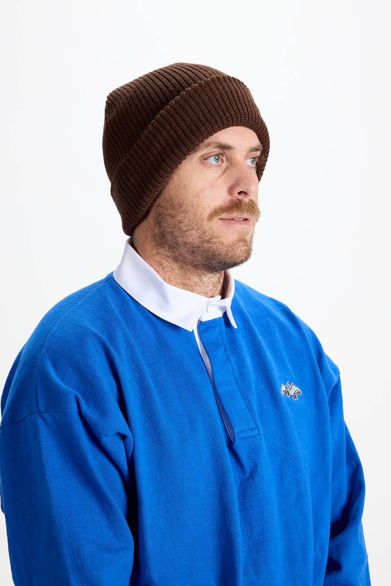 A man wearing a dark brown ribbed knit beanie and a blue half-zip rugby pullover over a white collared shirt, photographed against a plain white studio background.