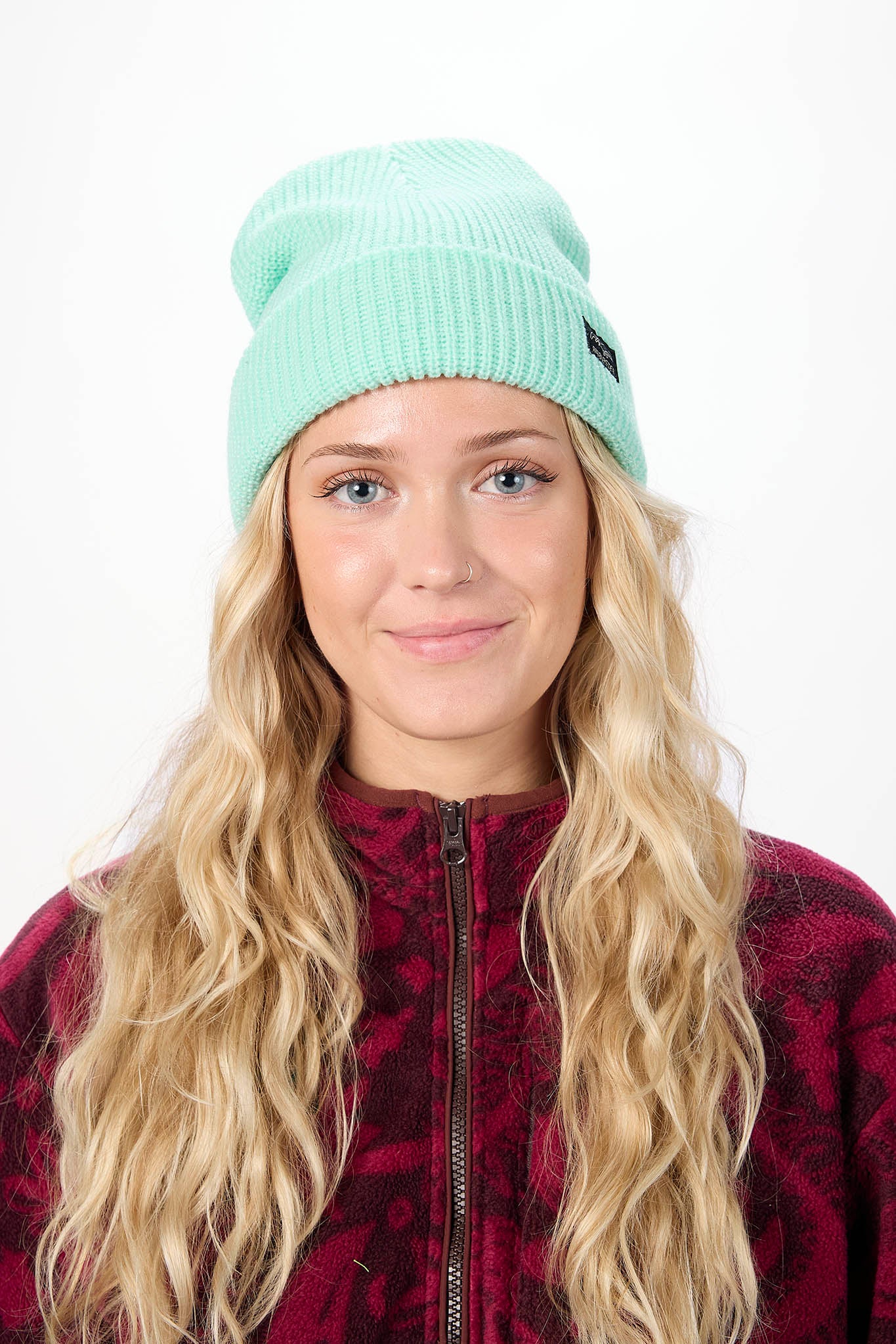 A woman with long blonde hair wearing a mint green ribbed knit beanie and a dark red patterned fleece jacket, photographed against a plain white background.