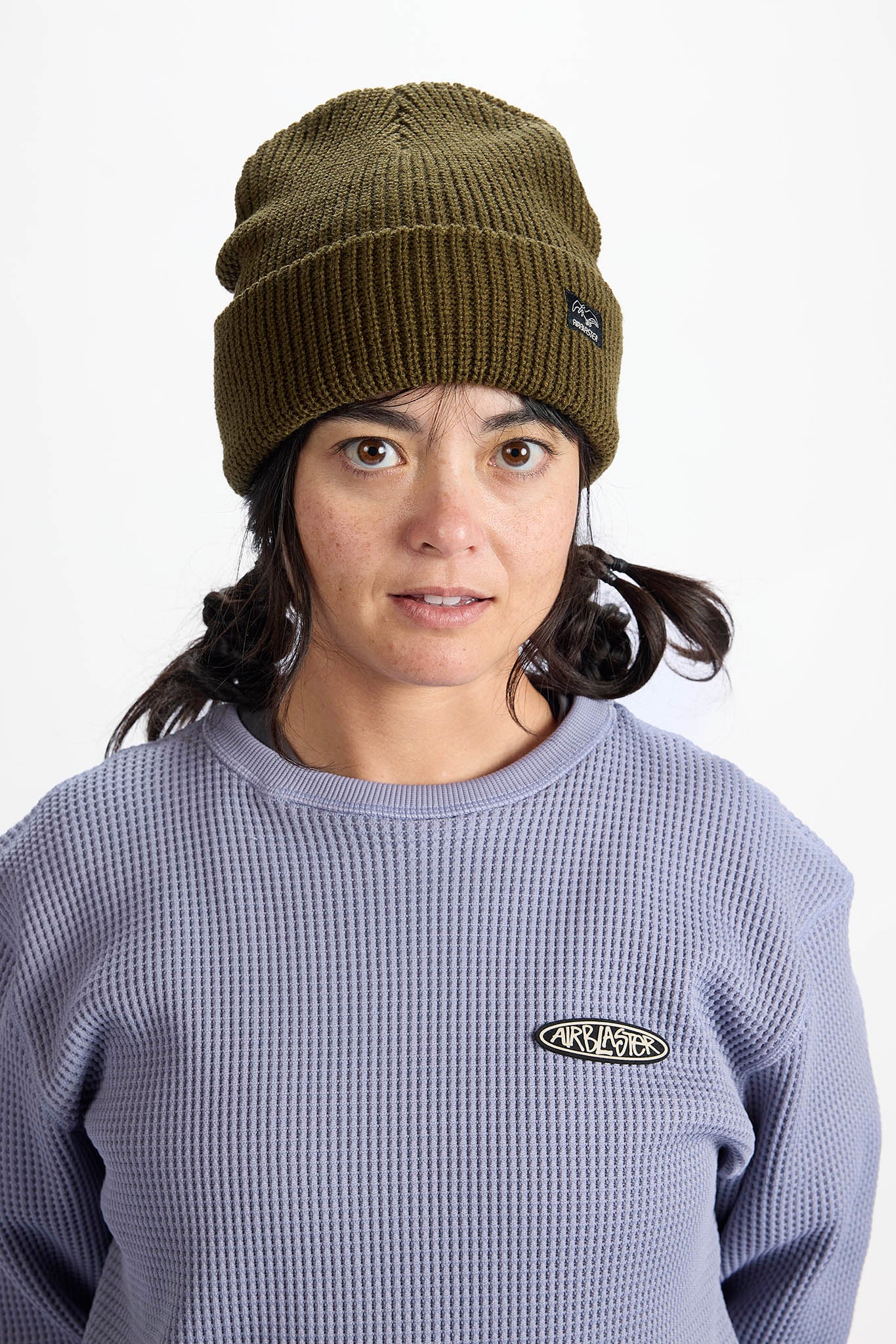 A woman wearing an olive green ribbed knit beanie and a lavender waffle-knit sweatshirt with an Anvil logo patch on the chest, photographed against a plain white studio background.