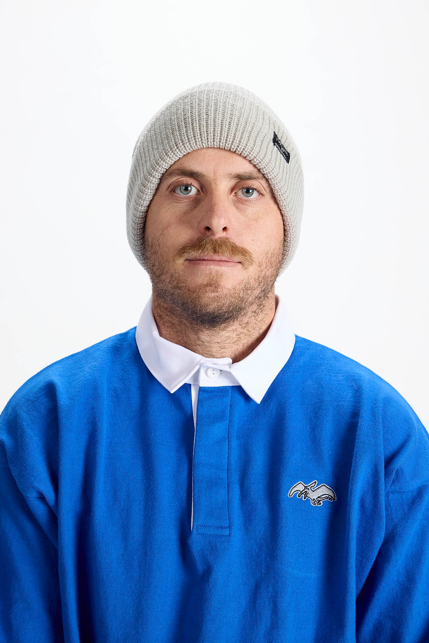 A man wearing a light gray ribbed knit beanie and a blue long-sleeve collared pullover sweater with a white collar, photographed against a plain white studio background.