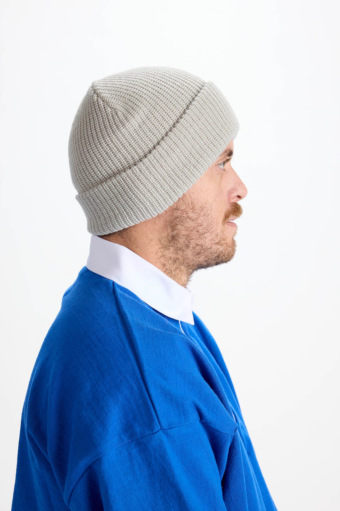 A man in profile wearing a light gray ribbed knit beanie, a blue sweatshirt, and a white collared shirt, photographed against a plain white studio background.