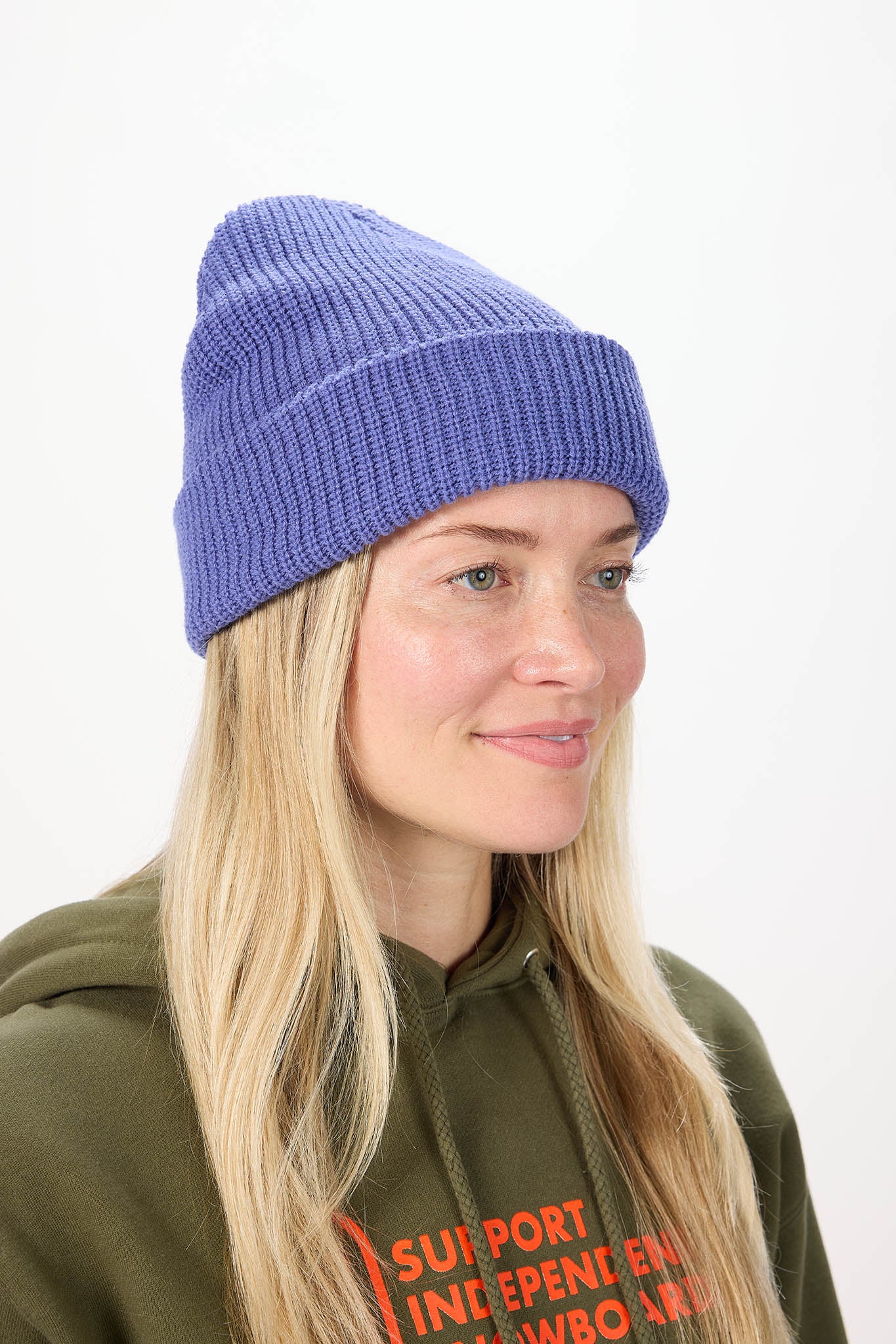 A woman with long blonde hair wearing a ribbed knit beanie in a periwinkle shade and an olive green hoodie with orange text, photographed against a plain white background.