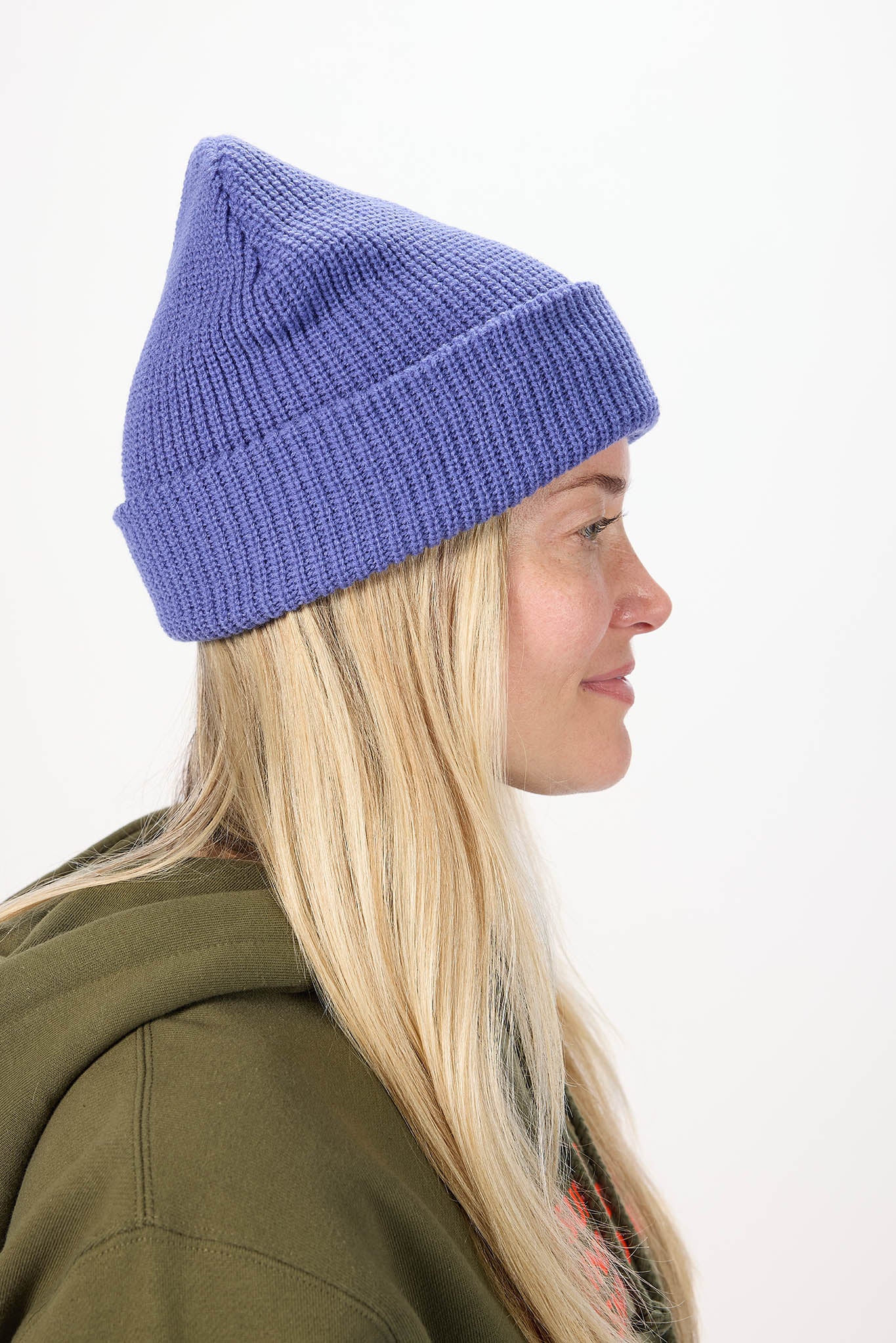 A woman in profile wearing a ribbed knit beanie hat in a periwinkle blue color, paired with an olive green hoodie, against a plain white background.