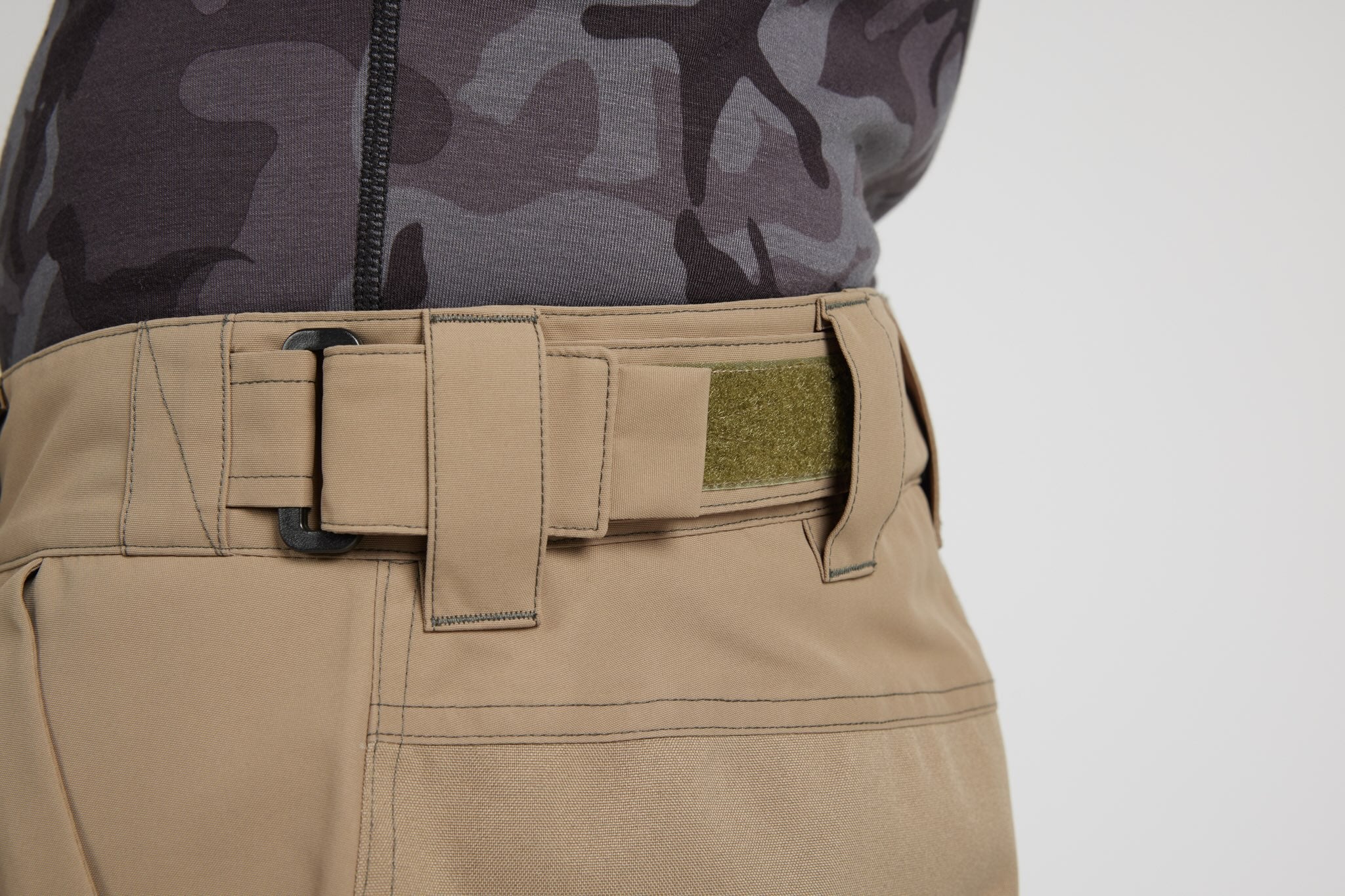 Close-up of tactical pants in khaki beige with a matching belt and a green hook-and-loop patch, worn with a dark gray camouflage top against a light gray background.