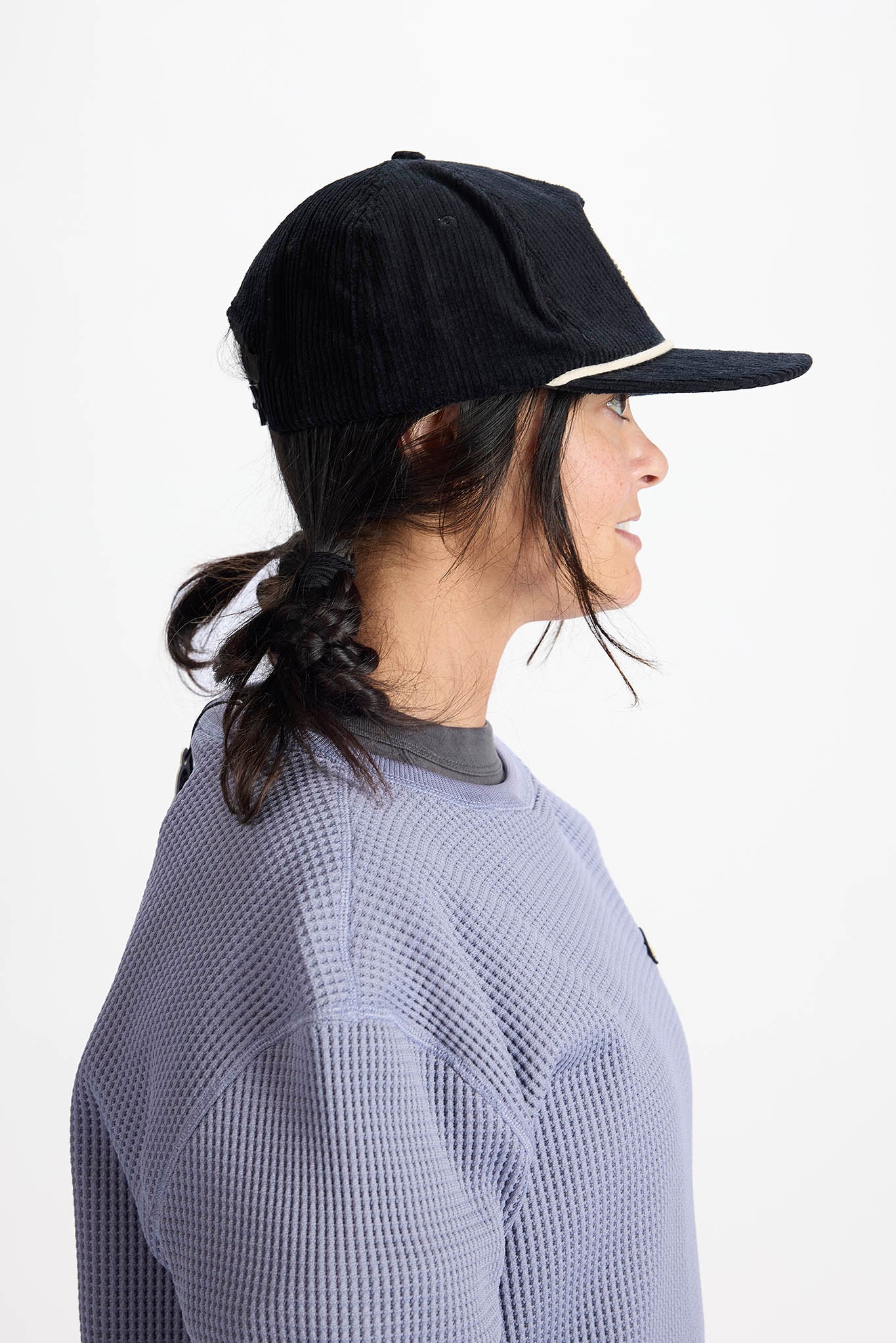 side profile A woman wearing a black Airblaster baseball cap and a lavender waffle-knit sweatshirt with a small Airblaster logo on the chest, photographed against a plain white background.
