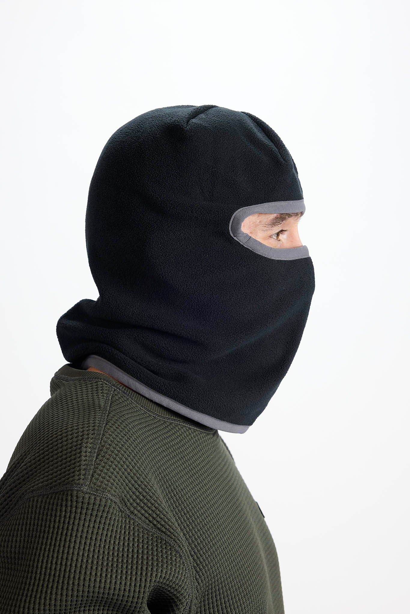 A person shown in side profile wearing a black balaclava mask with a gray trim around the eye opening, against a plain white background.