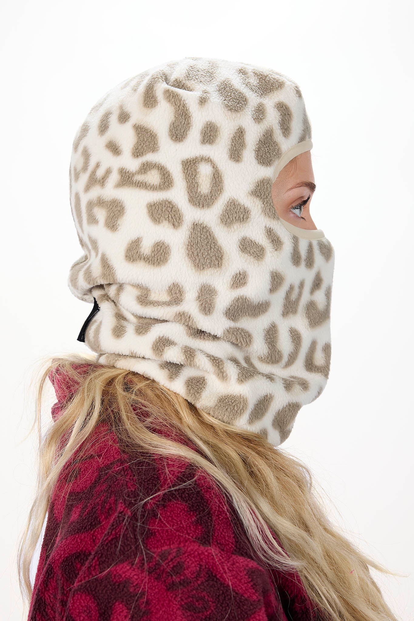 side profile A woman wearing a white leopard patterned balaclava mask with a brown leopard print pattern, paired with an olive green hoodie against a plain white background.
