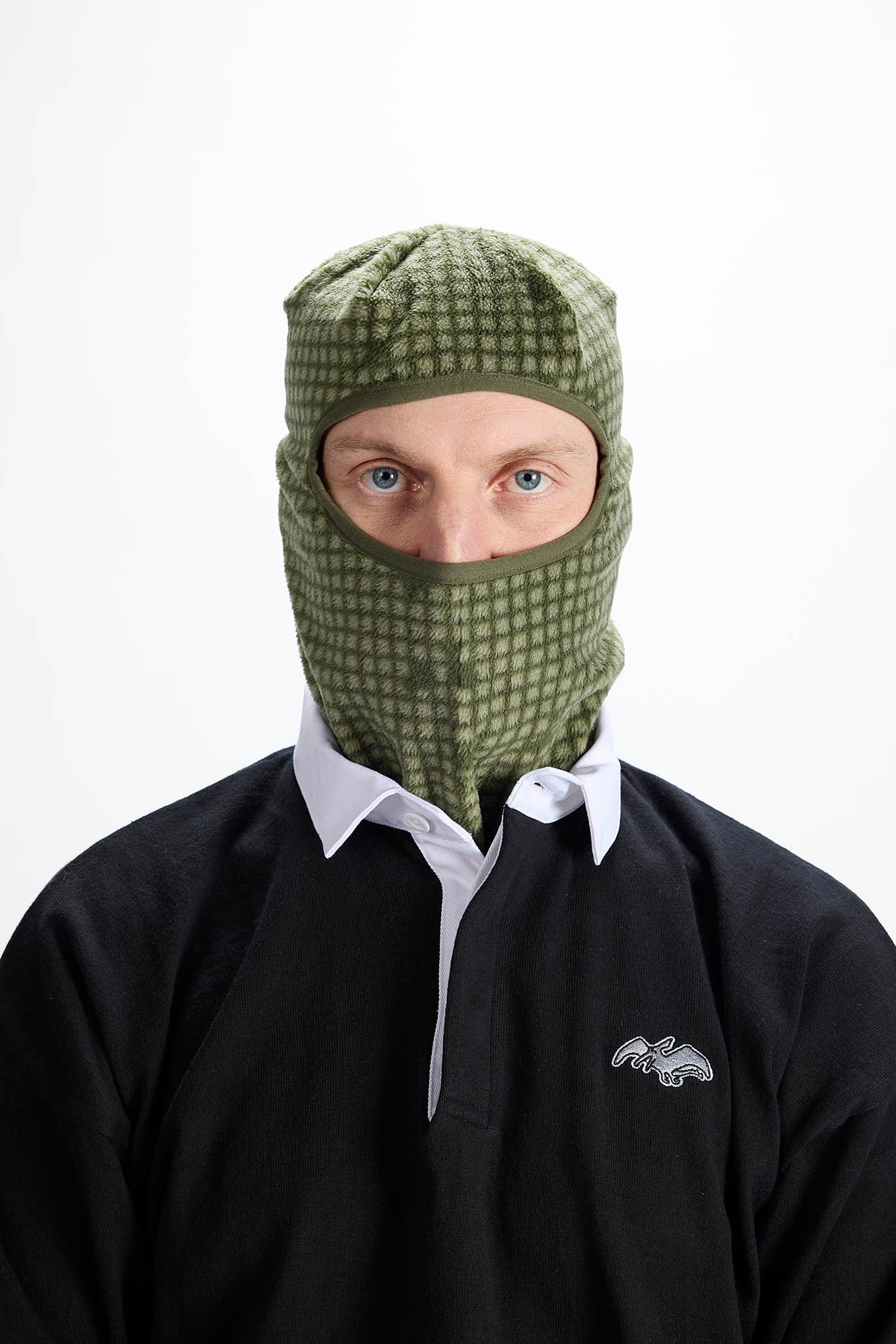 A man wearing an olive green and black checkered balaclava face covering over a black polo shirt with a white collar, photographed against a plain white background.