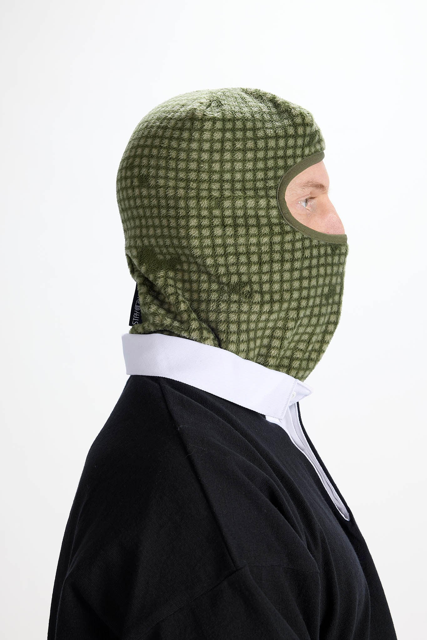 A person shown in side profile wearing an olive green knit balaclava mask with a white collared shirt underneath and a black jacket, against a plain white background.