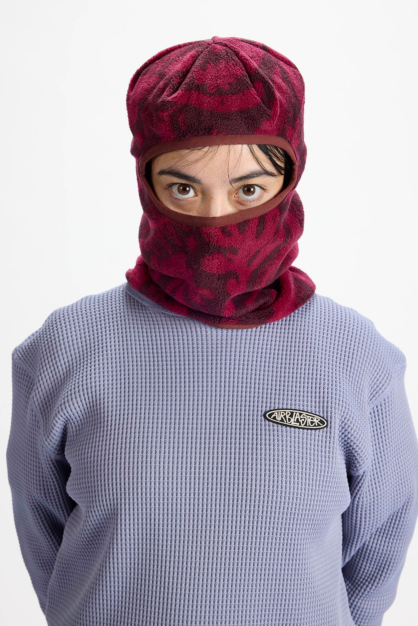 A model wearing a burgundy and black patterned fleece balaclava mask over their face, paired with a lavender waffle-knit sweatshirt featuring a small oval logo on the chest.