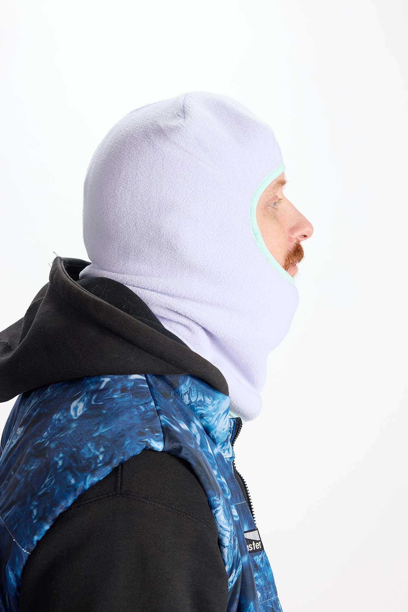 in side profile A man wearing a light purple balaclava mask with a brown leopard print pattern, paired with an olive green hoodie against a plain white background.