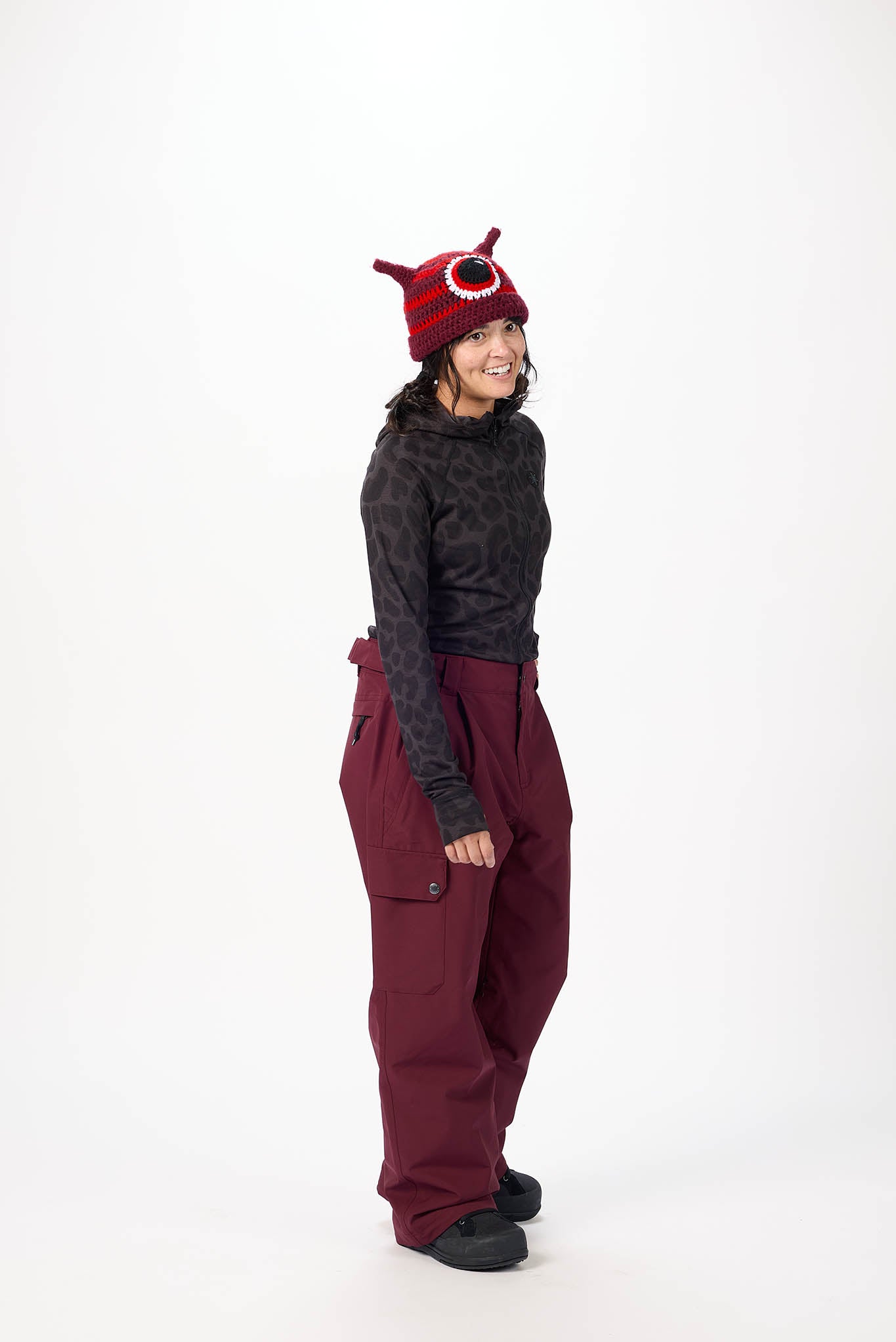 Person wearing a dark long-sleeve shirt, maroon pants, and a red and black beanie with horns on a white background