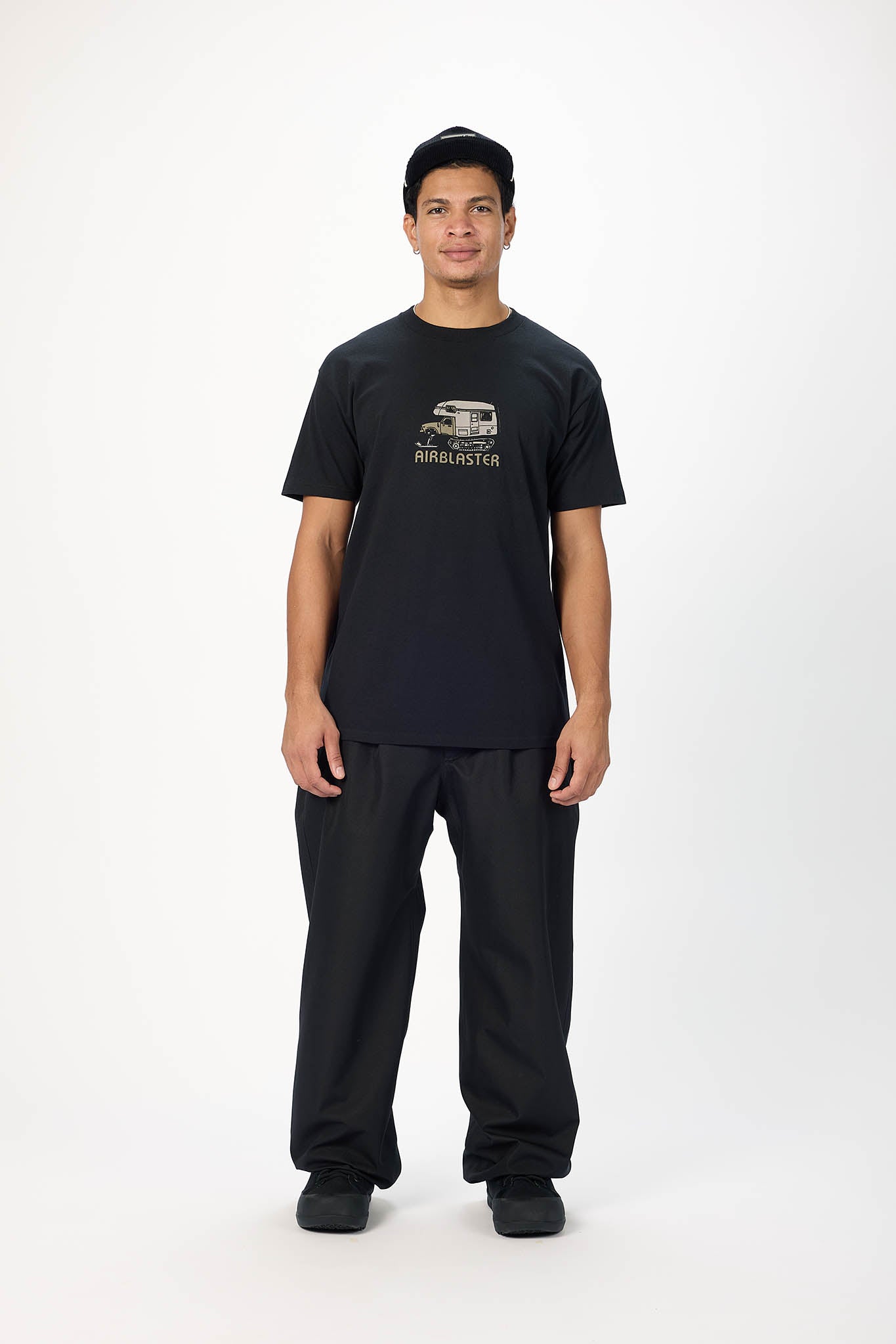 A young man modeling a black crewneck t-shirt with a vintage-style graphic and text on the chest, paired with black pants, against a plain white studio background.