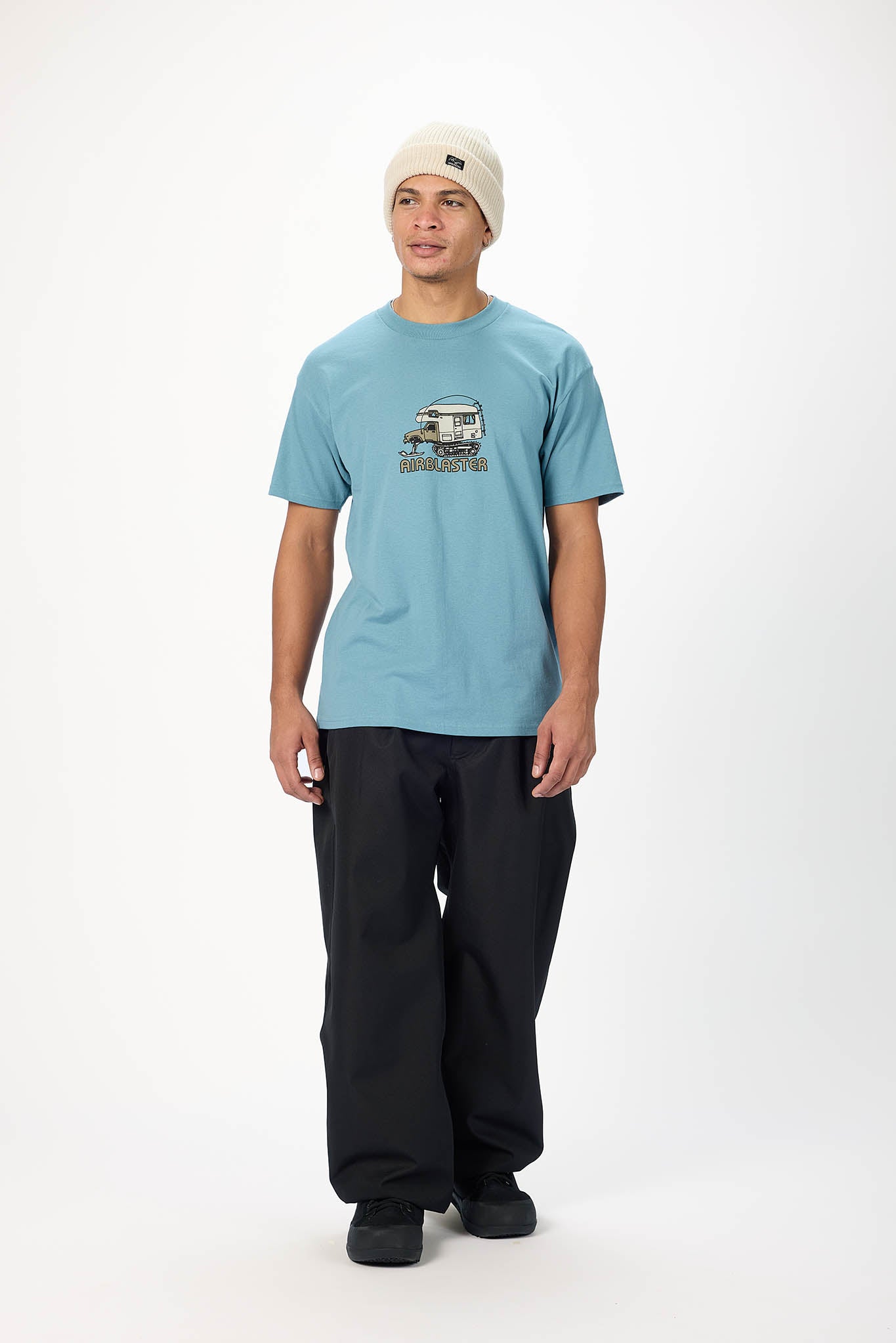 A man wearing a light blue graphic t-shirt, black pants, and a cream beanie stands against a plain white studio background.