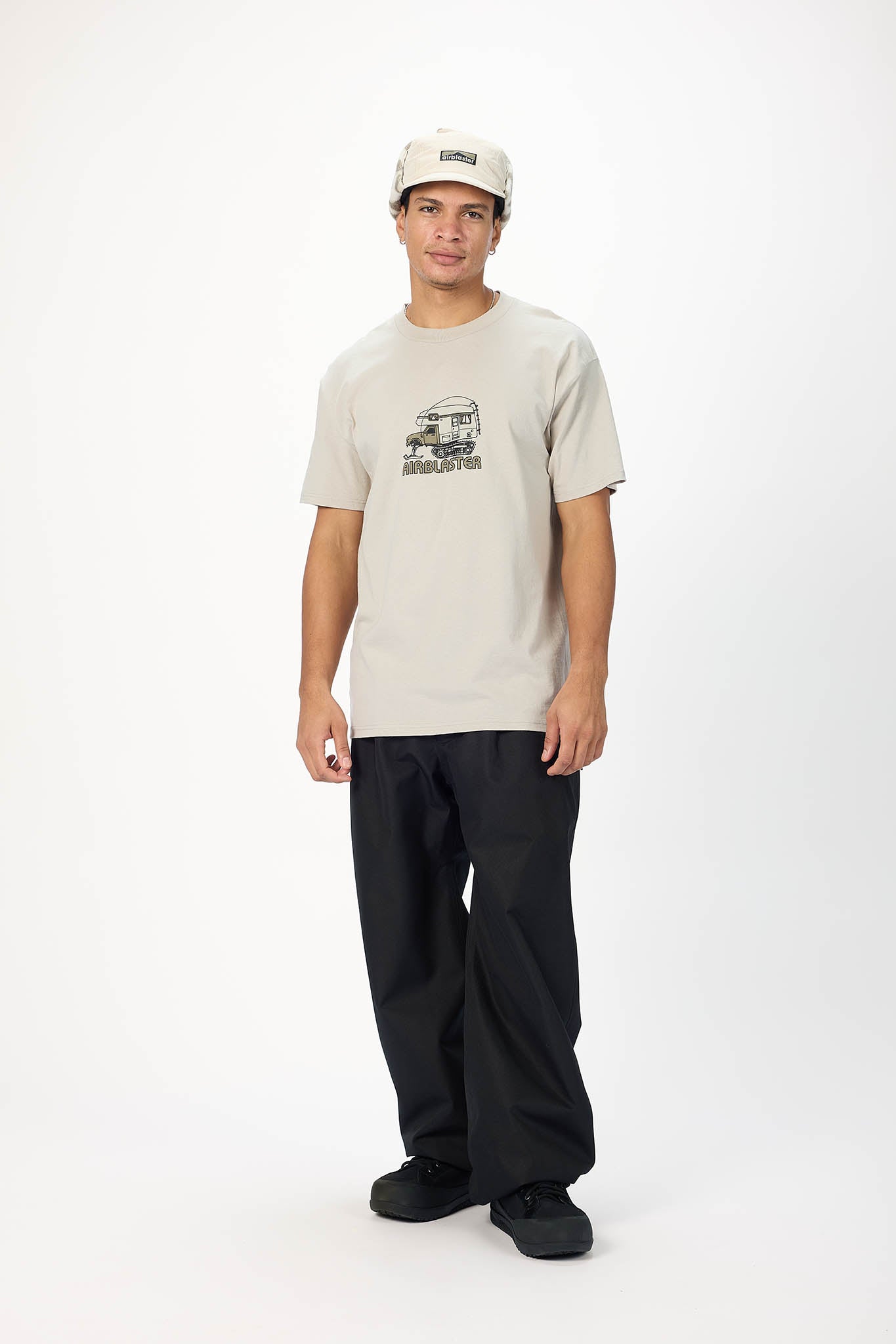 A man modeling a light beige graphic t-shirt, black pants, and a beige baseball cap against a plain white studio background.