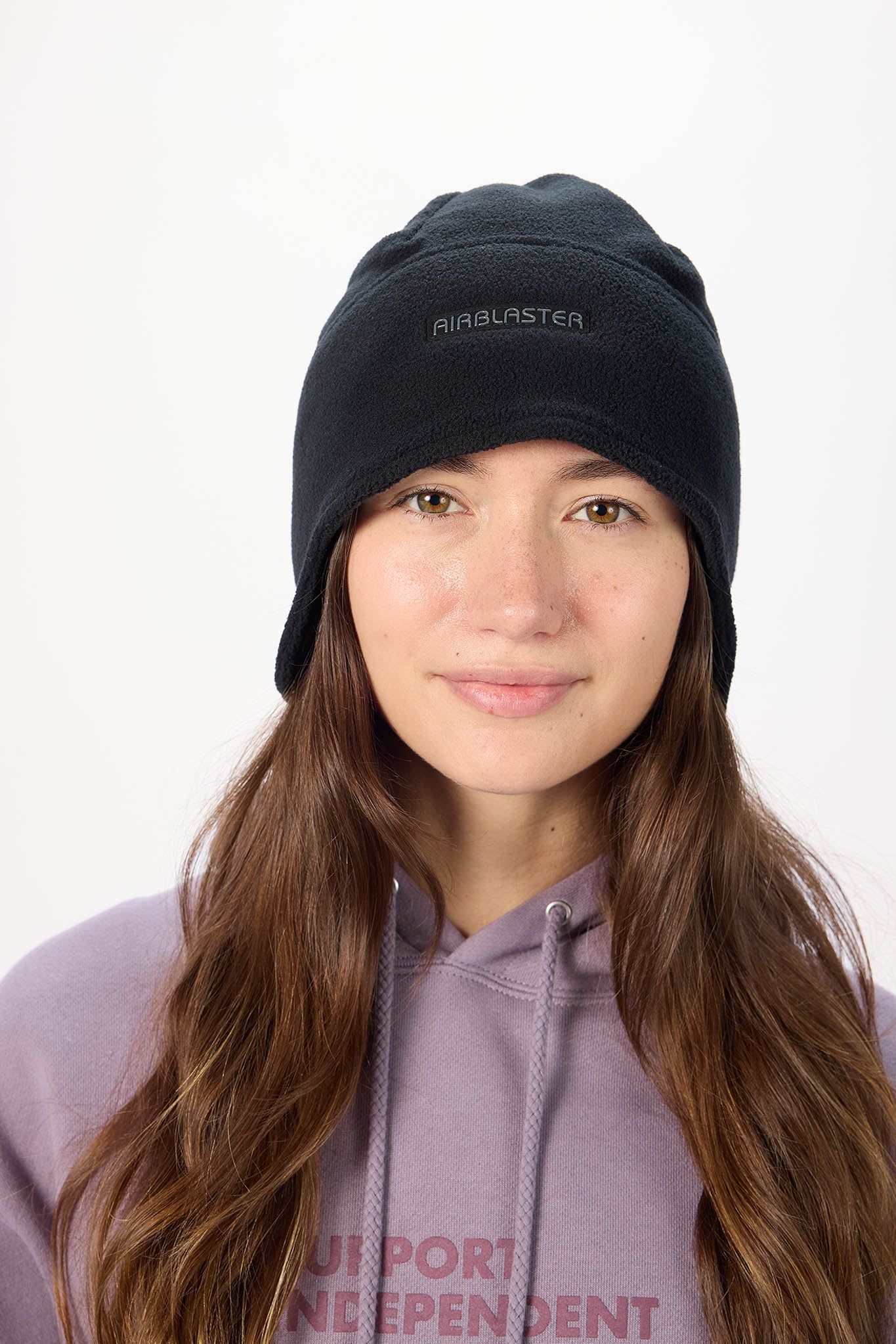 A woman with long brown hair wearing a black knit beanie with embroidered in white on the front, paired with a lavender hoodie against a plain white background.