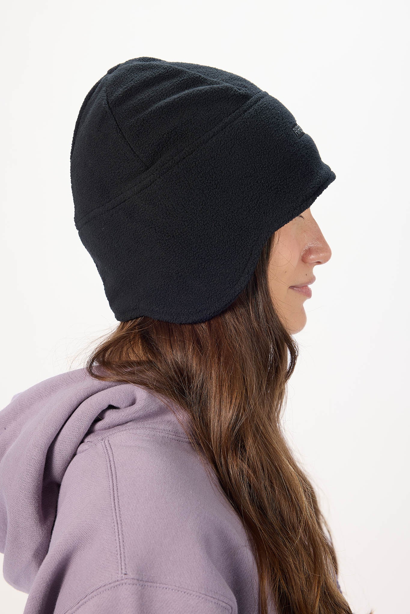 A woman in profile wearing a black slouchy knit beanie and a lavender hoodie, photographed against a clean white background.