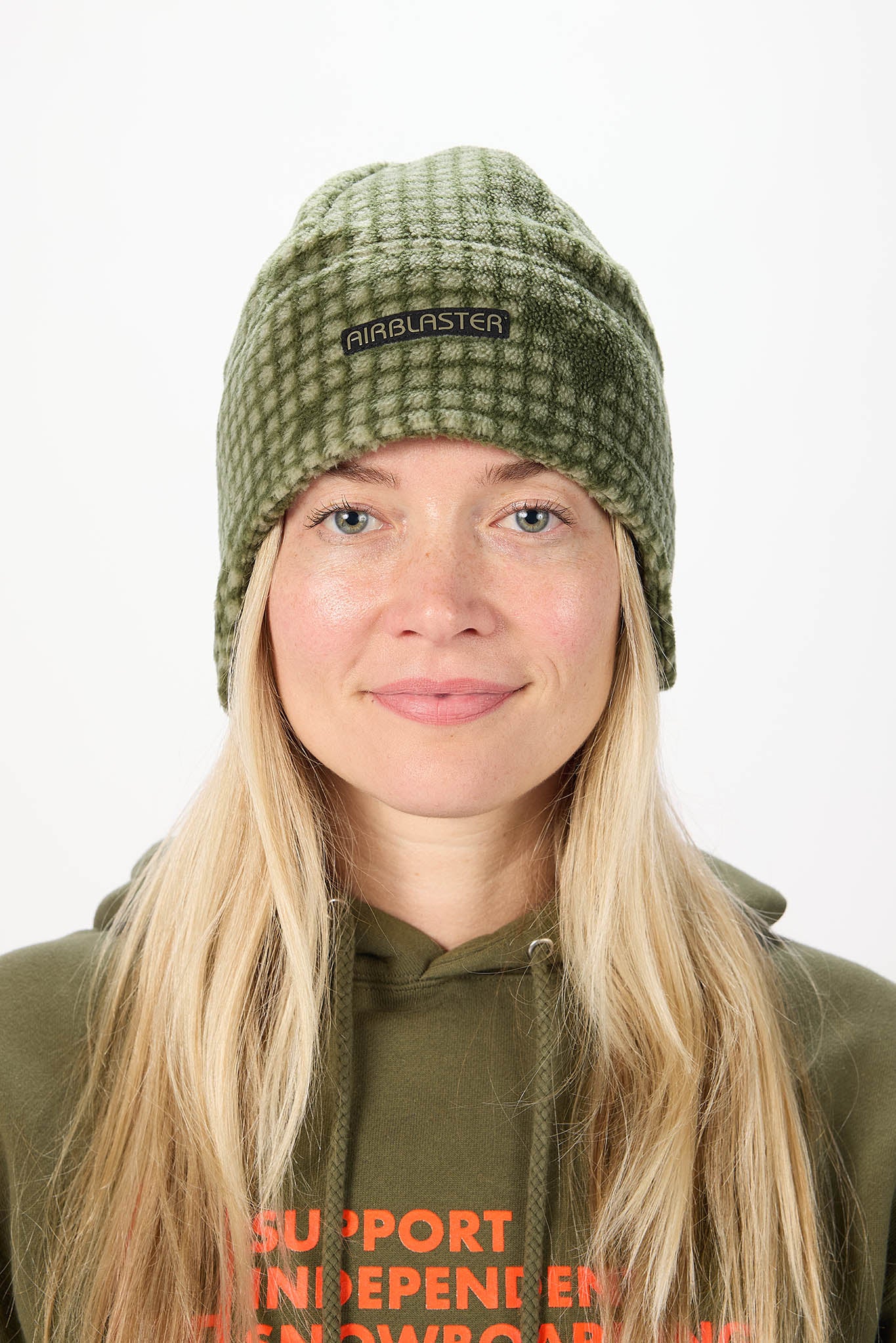 A woman with long blonde hair wearing an olive green checkered knit beanie with a black 'airblaster' logo patch, paired with an olive green hoodie against a plain white background.