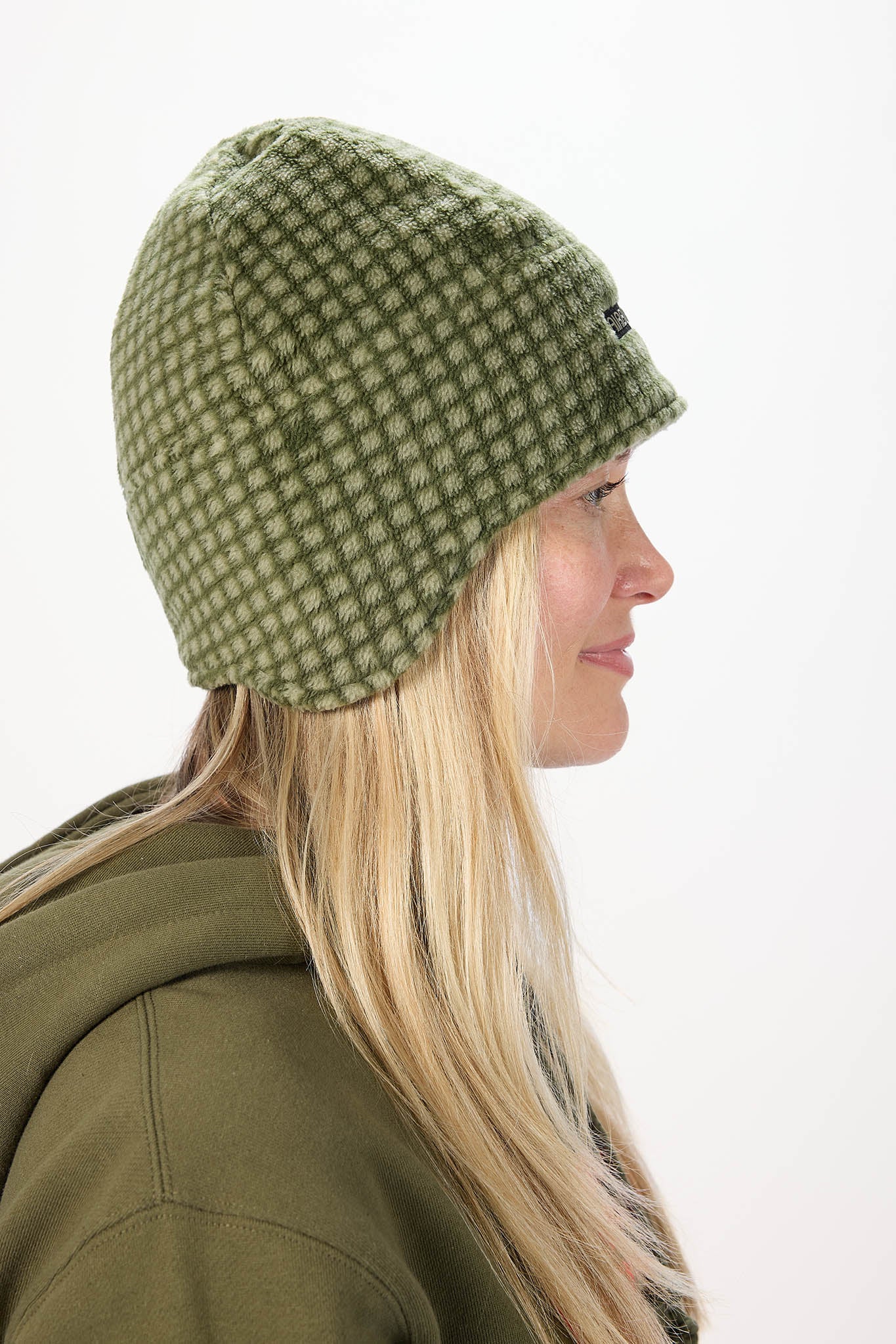 A woman in profile wearing an olive green and white checkered knit beanie hat and an olive green hoodie, set against a plain white background.