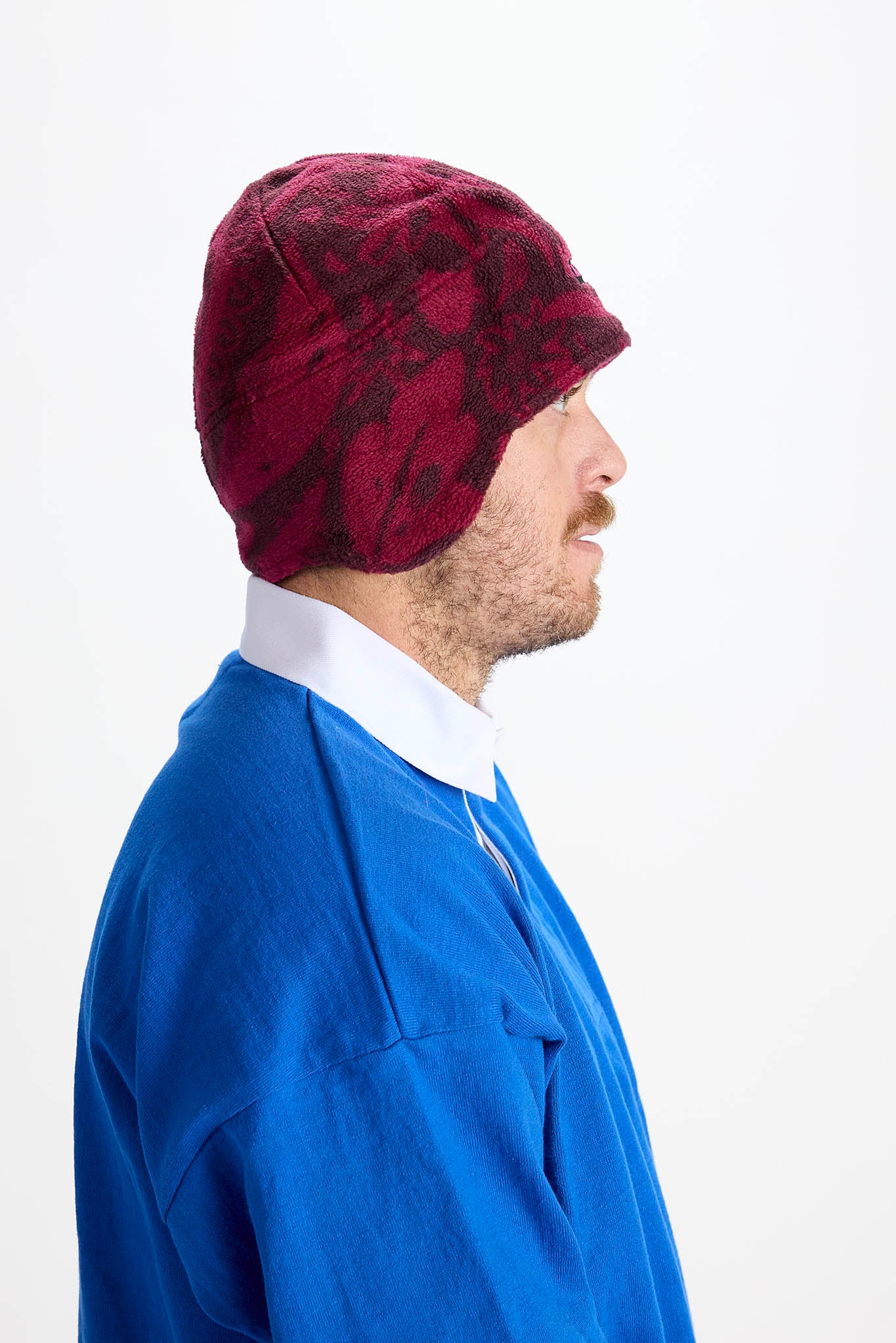 A man in profile wearing a burgundy and black patterned knit beanie with ear flaps, a blue shirt, and a white collared shirt, against a plain white background.