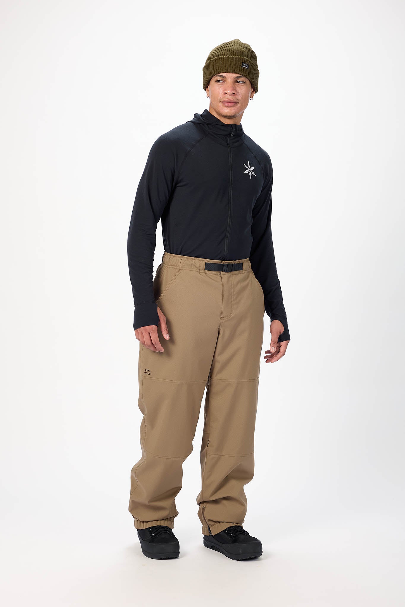Person wearing a black jacket with a logo, beige pants, and a green beanie on a white background