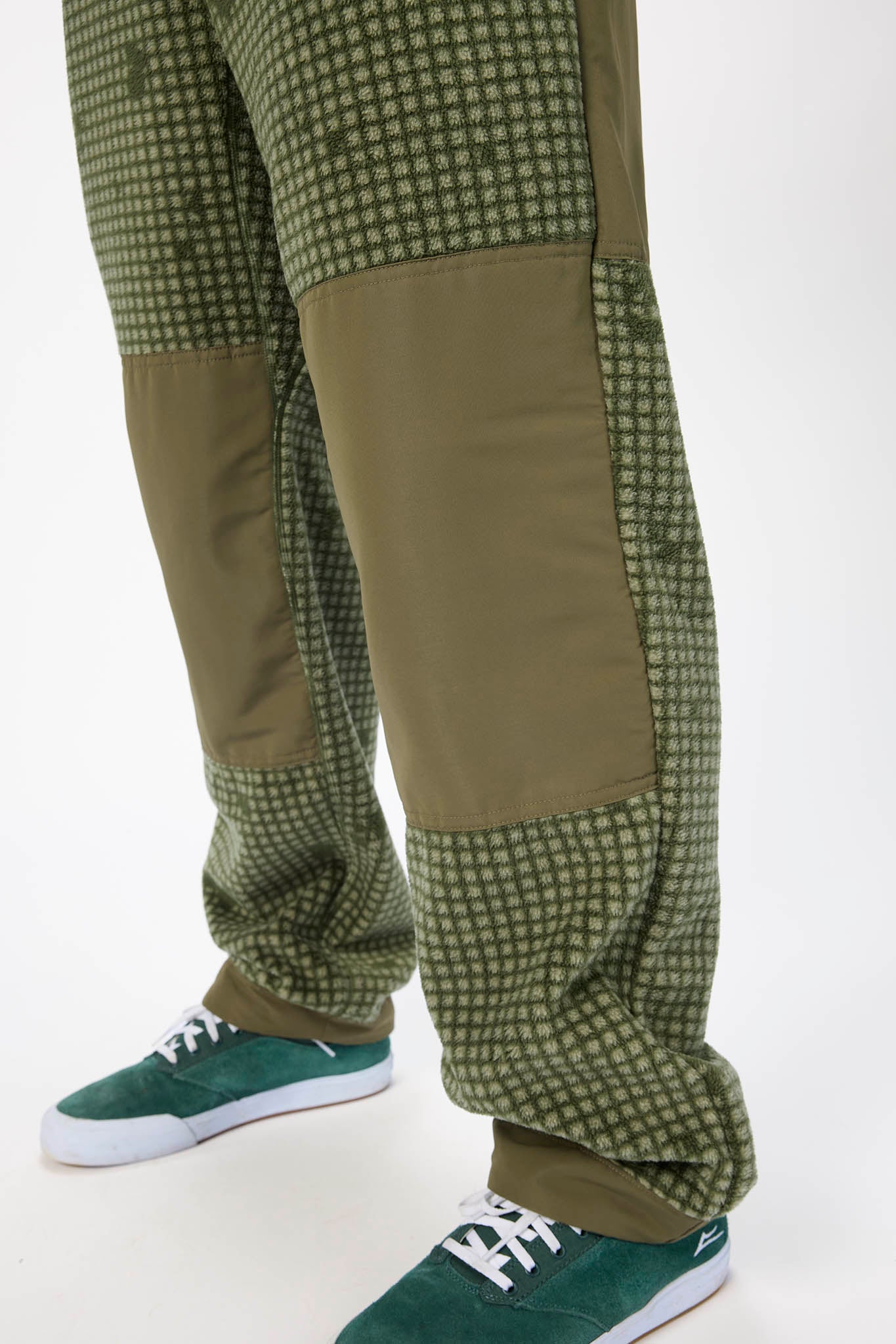 Close-up of a person's legs wearing olive green patchwork pants with a small checkered pattern and solid olive knee panels, paired with green suede sneakers.