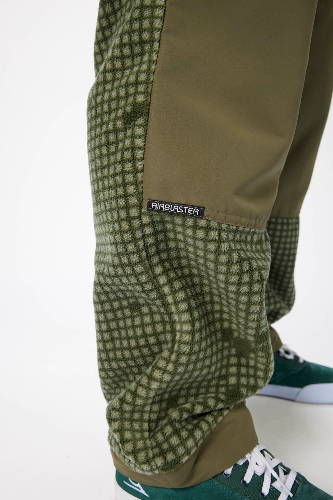 Close-up of a person's lower legs wearing olive green and black checkered fleece pants with a solid olive green panel, paired with green sneakers.