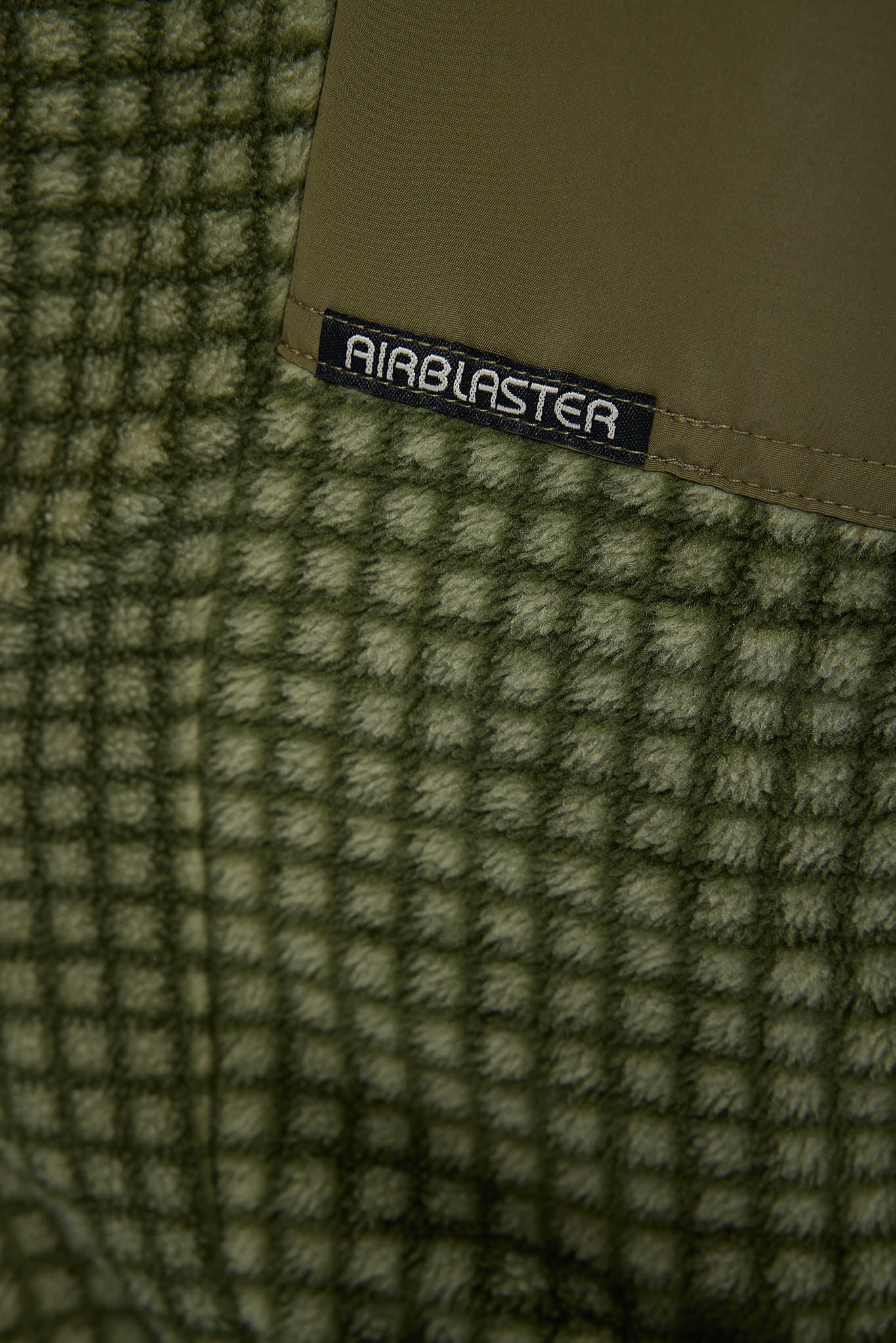 Close-up of a textured knit fabric in olive green and beige with a tan patch and a black AIRBLASTER brand label stitched onto it.
