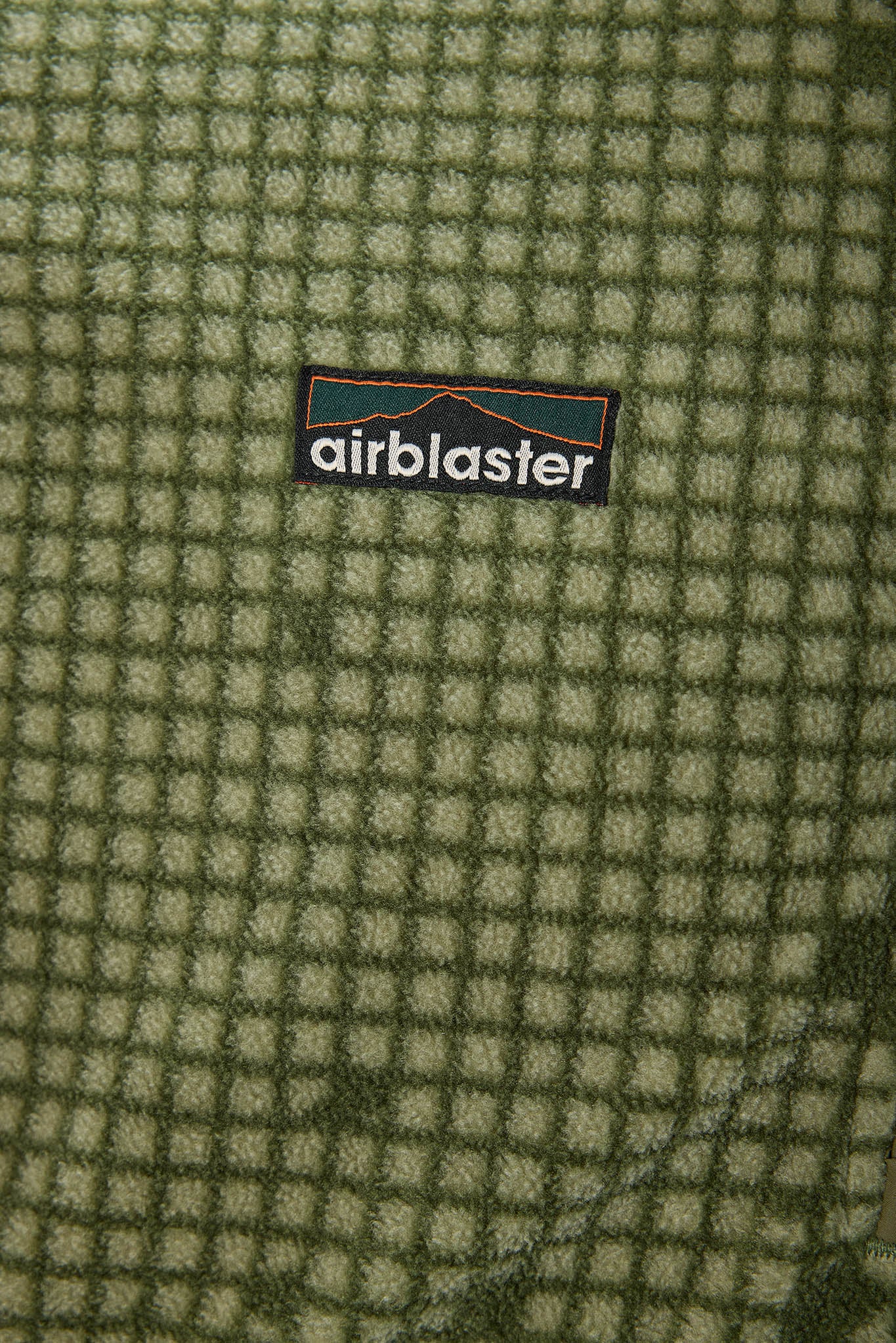 Close-up of a textured fleece fabric with a green and beige grid pattern, featuring a central black Airblaster logo patch.