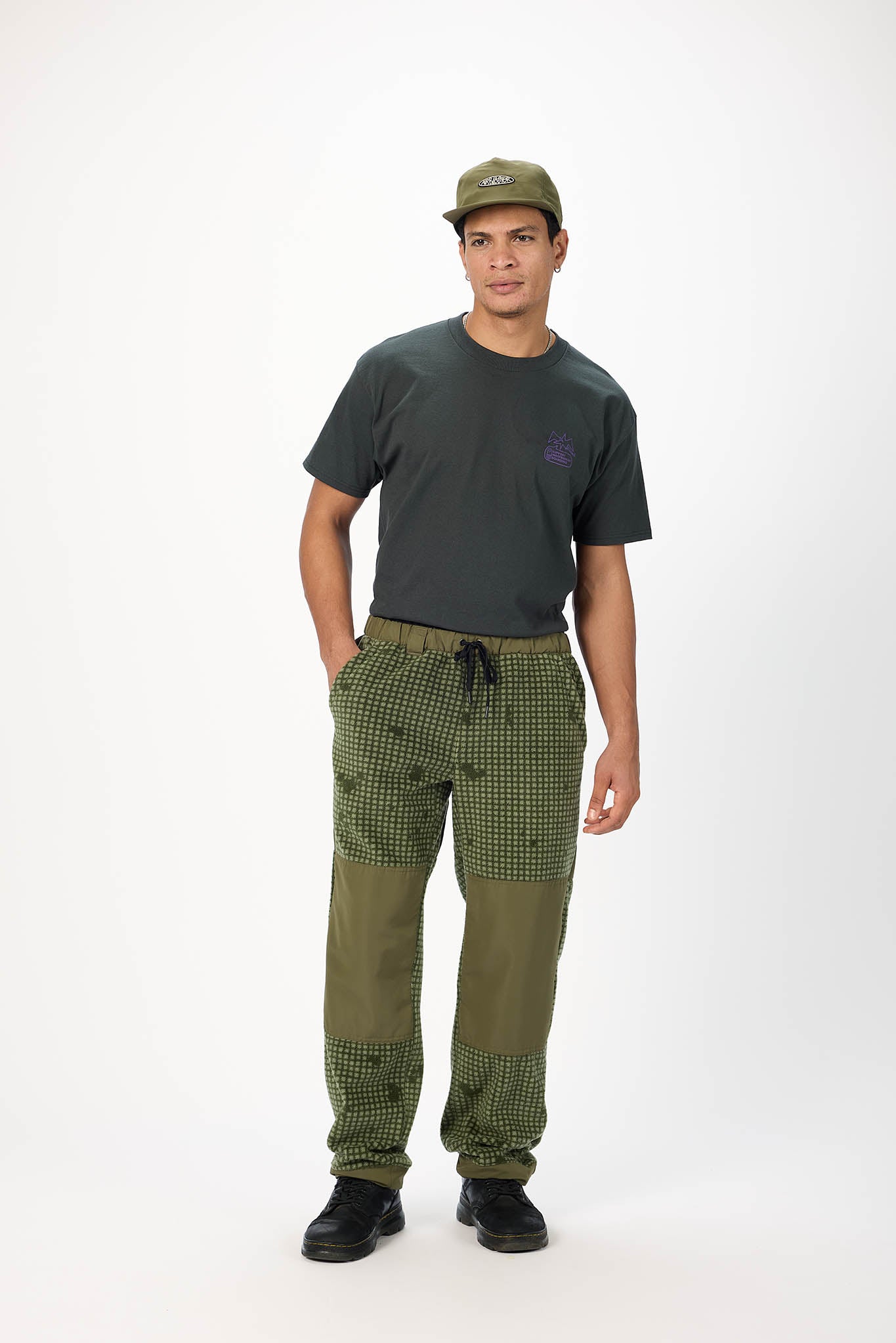 A man modeling a dark green crewneck t-shirt, olive green patchwork cargo pants, and an olive green baseball cap against a plain white studio background.