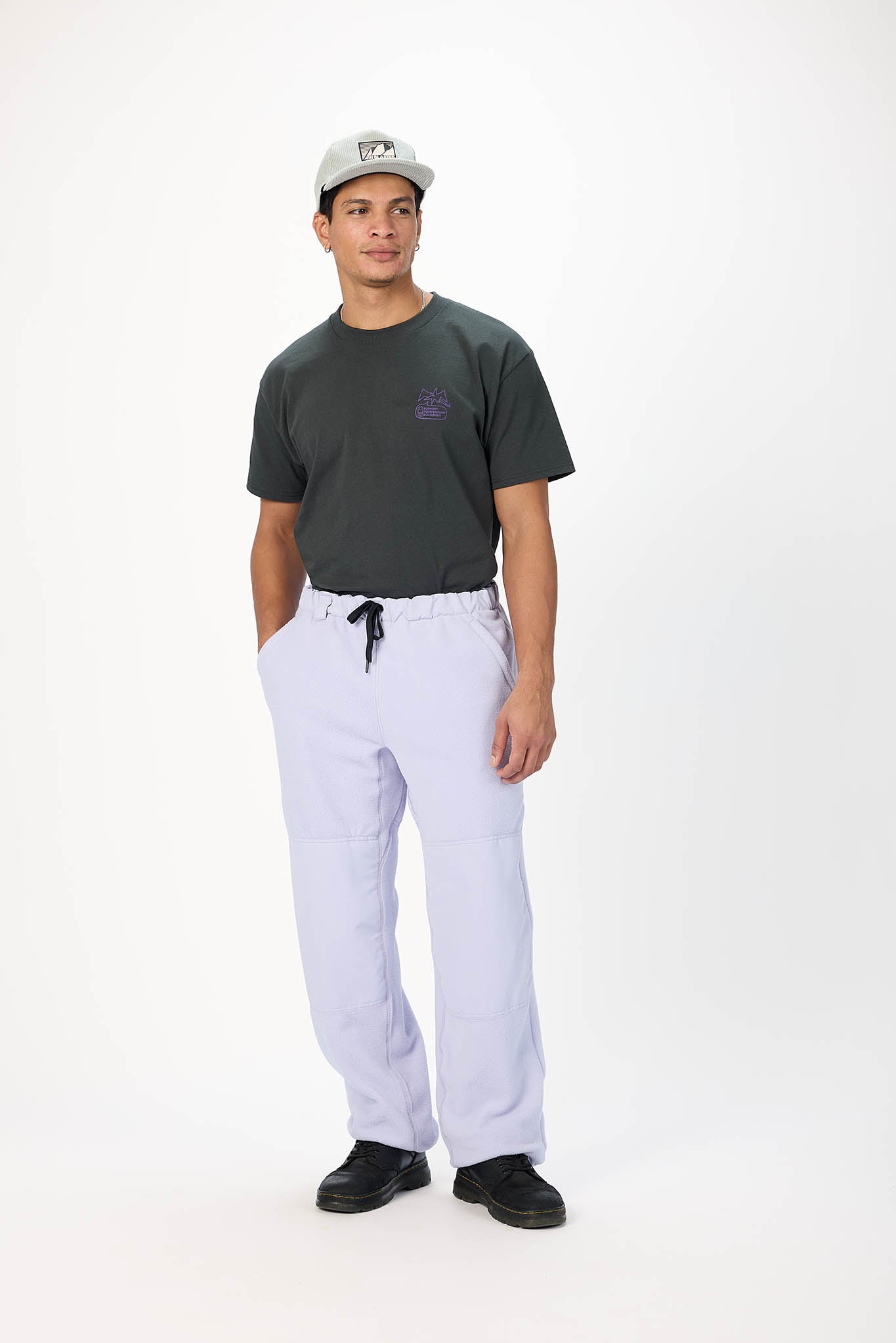 A man modeling a dark charcoal t-shirt, light lavender drawstring sweatpants, and a gray baseball cap against a plain white studio background.