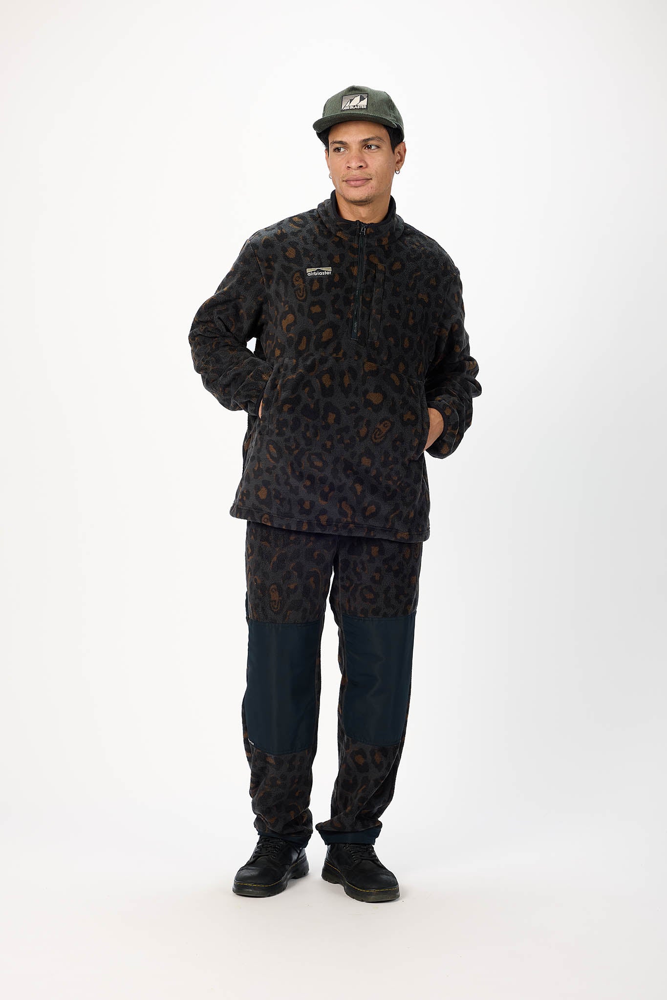 A man modeling a matching dark patterned fleece jacket and pants set with dark blue jeans, black boots, and a dark green baseball cap against a white studio background.