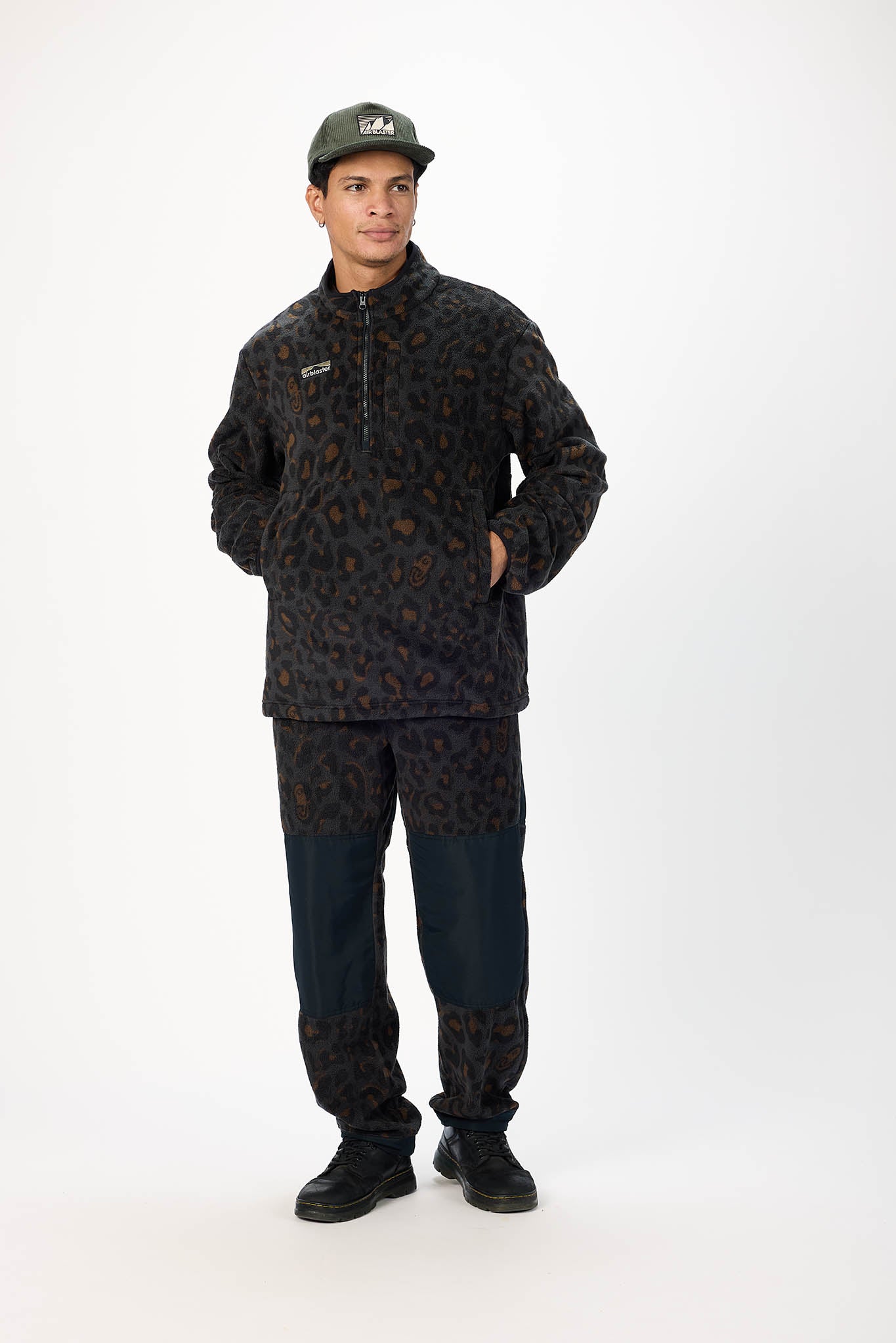 A man modeling a dark patterned fleece jacket and matching pants with dark navy lower legs, paired with a dark green cap, against a plain white studio background.