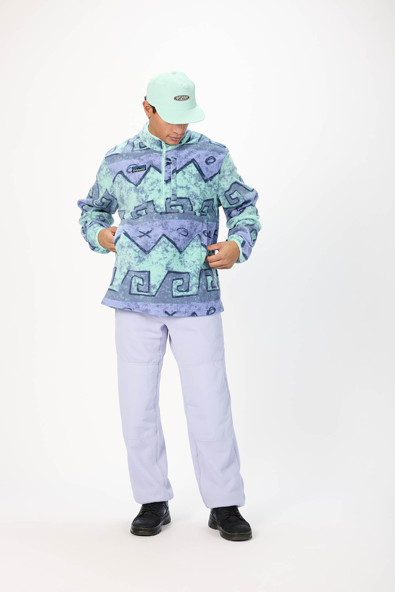 A man modeling a light blue and purple patterned quarter-zip pullover hoodie, paired with light purple pants, black shoes, and a mint green cap, against a plain white studio background.