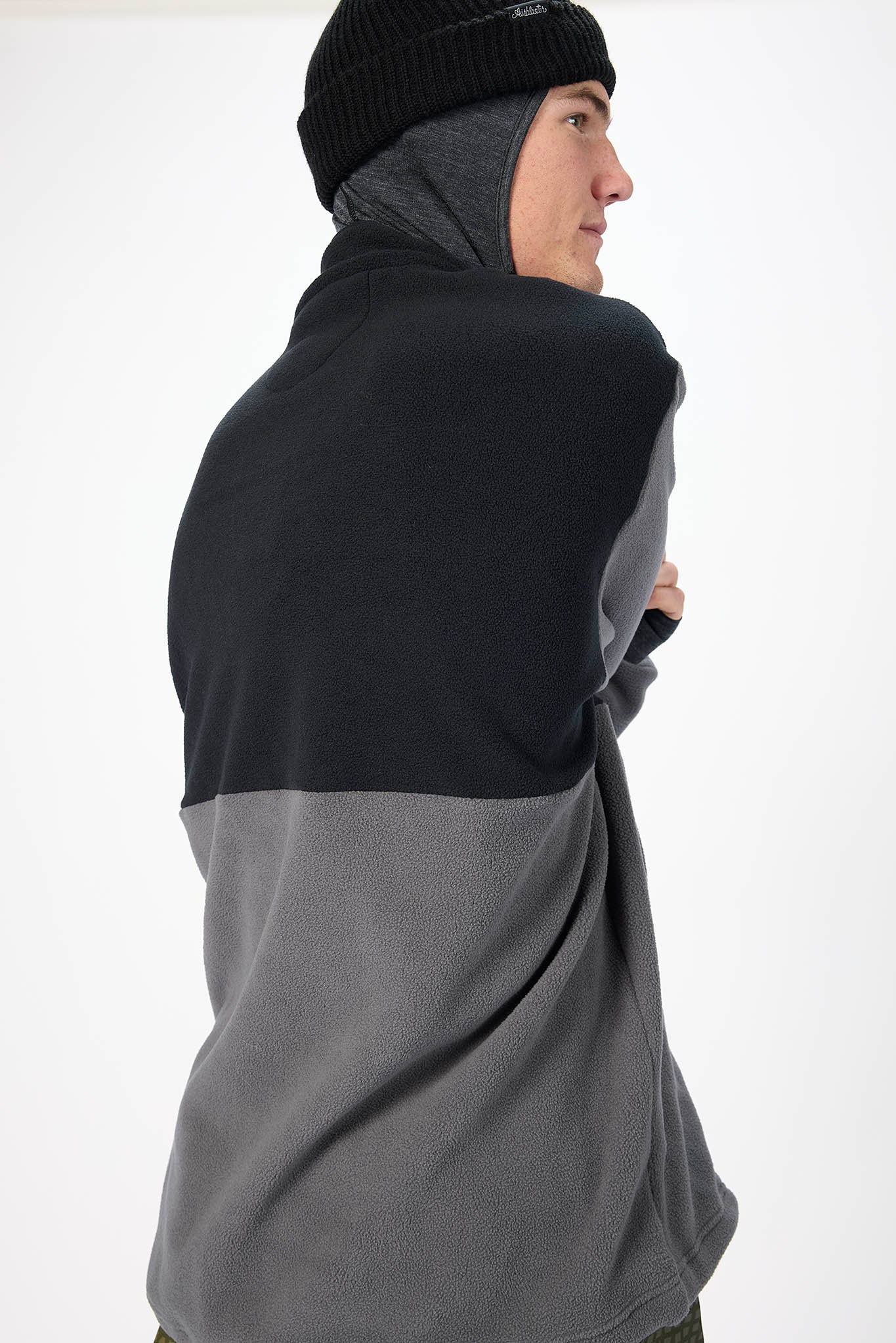 A man wearing a colorblock fleece hoodie with a black upper section and gray lower section, paired with a black beanie, shown from a side-back profile against a white studio background.