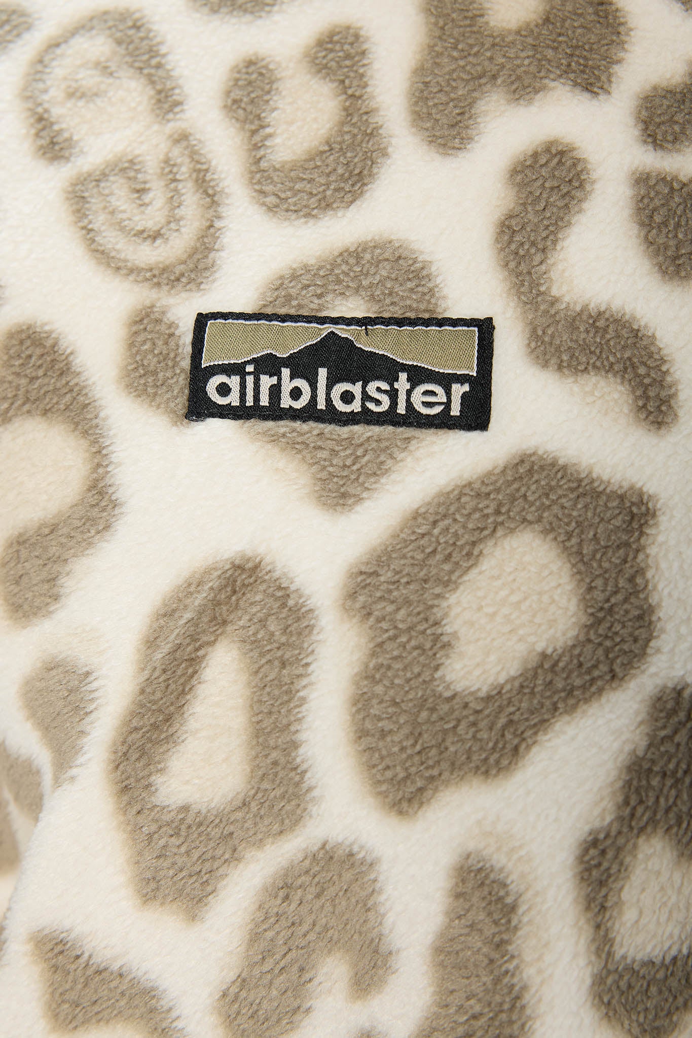 Close-up of a plush fleece fabric with an animal print pattern in beige and brown on an off-white background, featuring a black Airblaster logo patch.