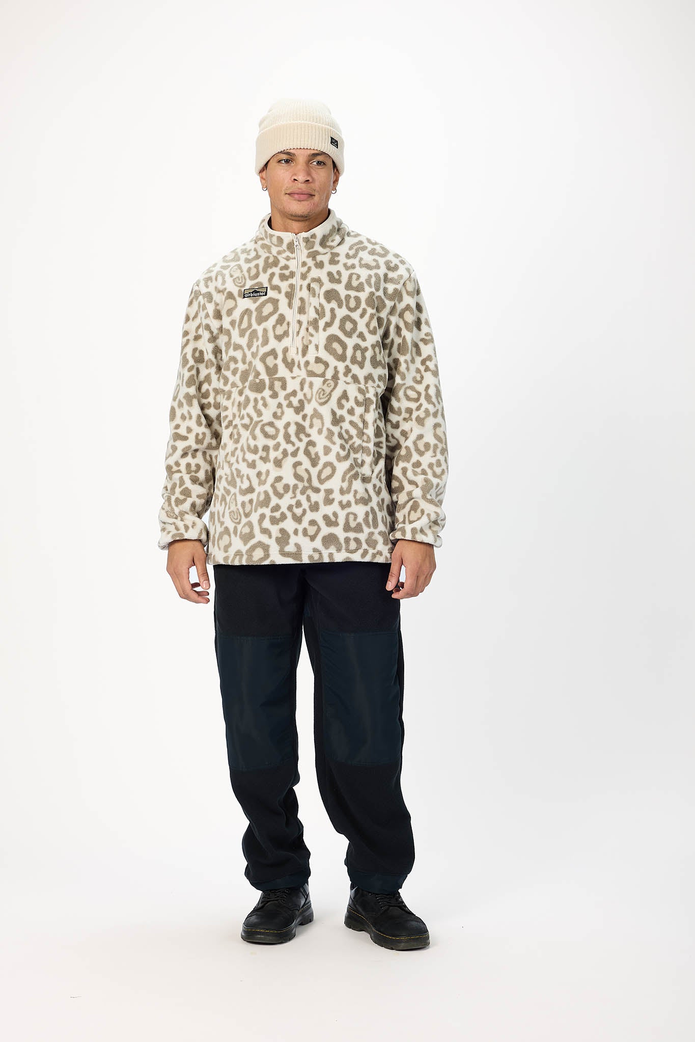 A man modeling a beige and brown leopard-print quarter-zip pullover jacket, paired with dark pants and a cream beanie, standing against a plain white studio background.