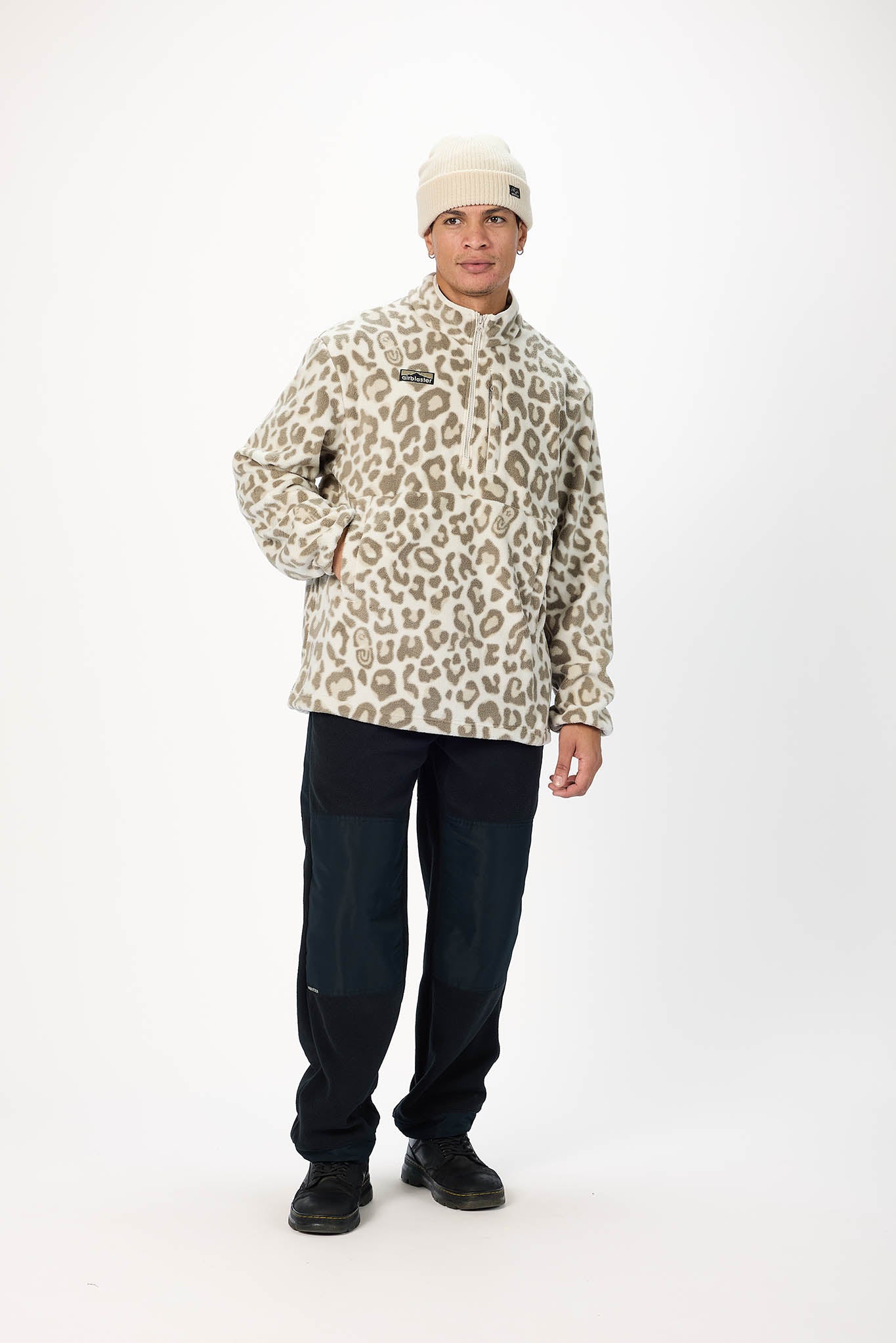 A man modeling a beige and brown leopard-print fleece jacket, paired with dark pants and a cream beanie, standing against a plain white studio background.