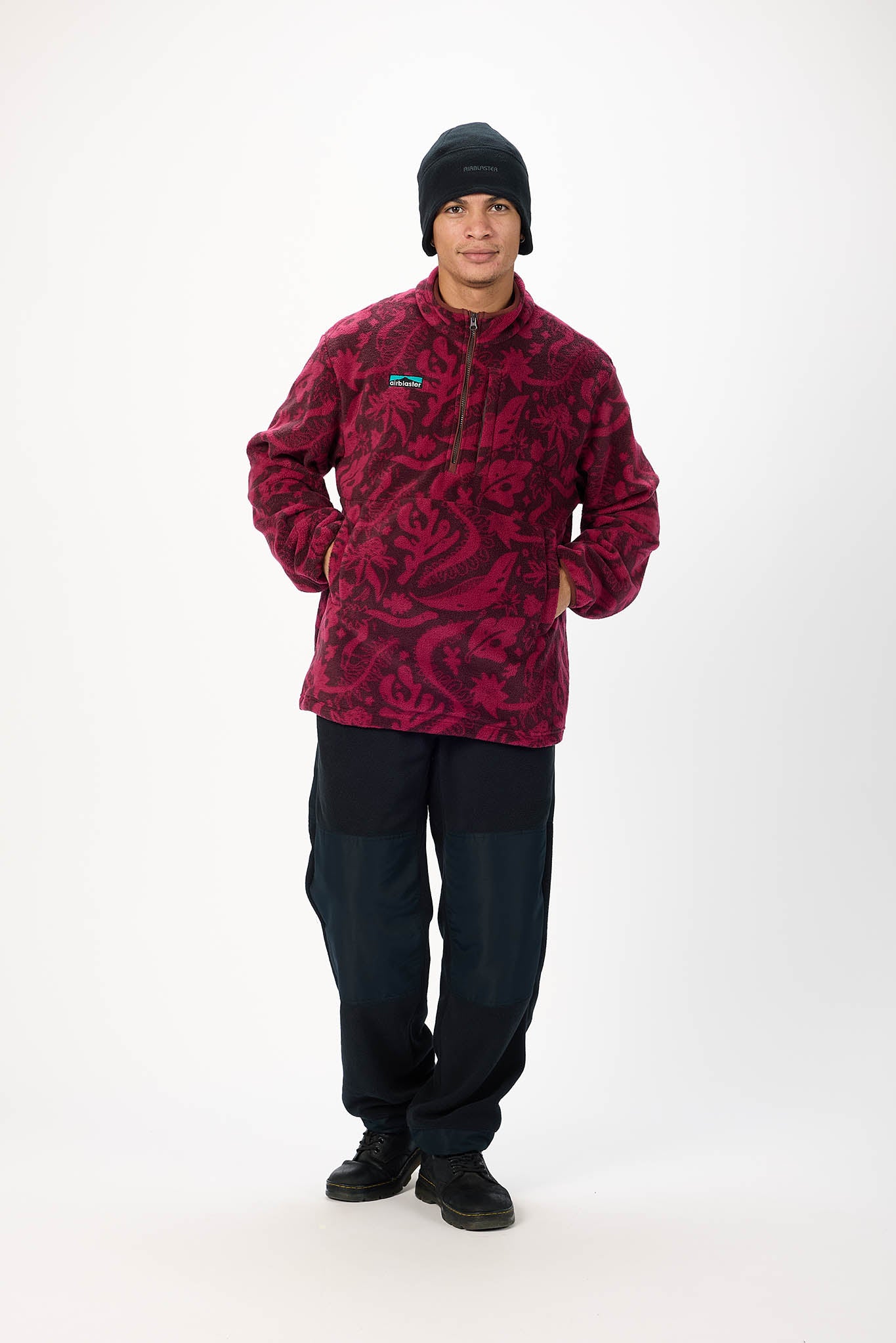 A man modeling a fleece pullover jacket with a dark red and black abstract pattern, paired with dark pants, black shoes, and a black beanie, against a plain white studio background.