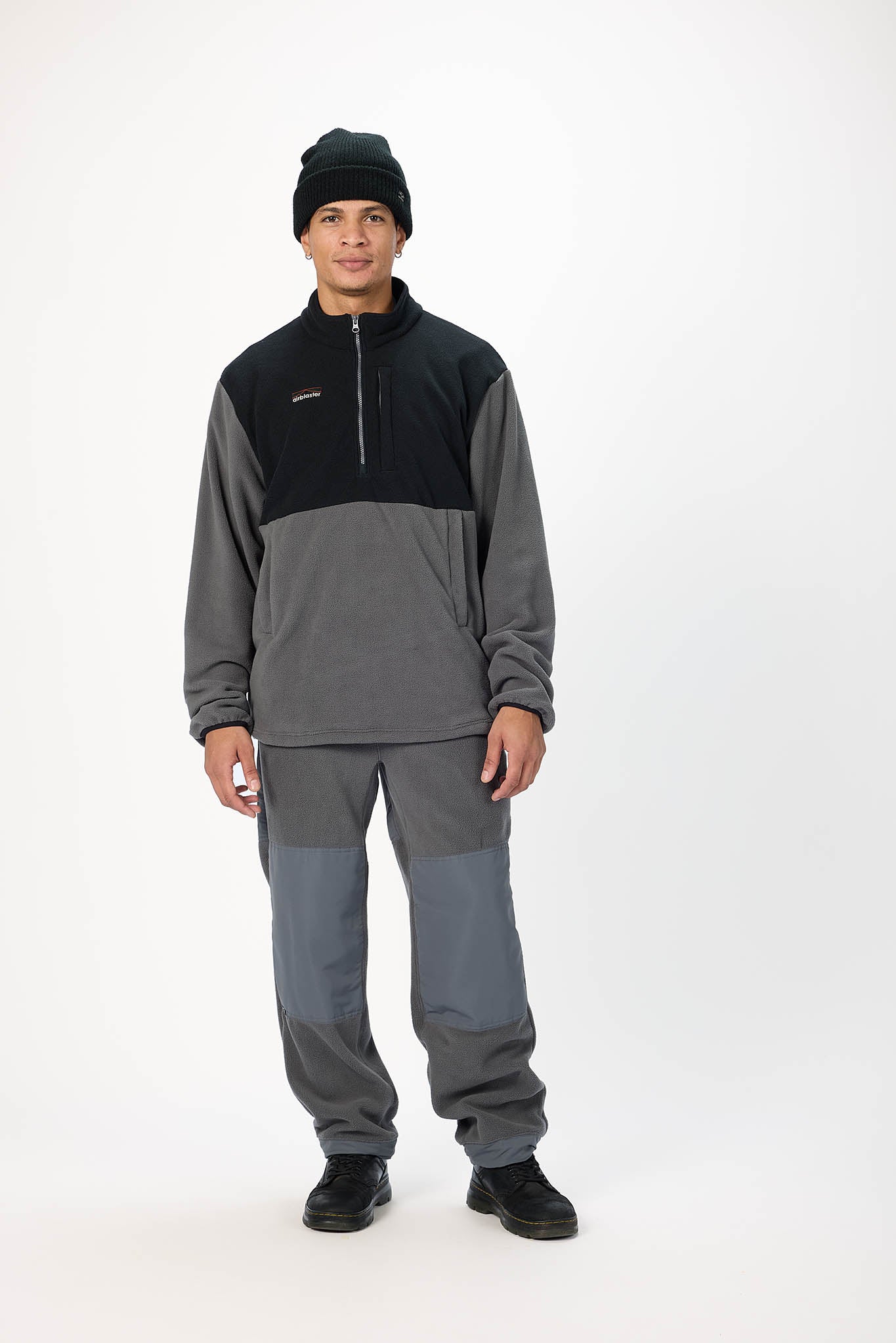 A man modeling a two-tone gray and black quarter-zip fleece pullover with matching gray pants and a black beanie, standing against a plain white studio background.