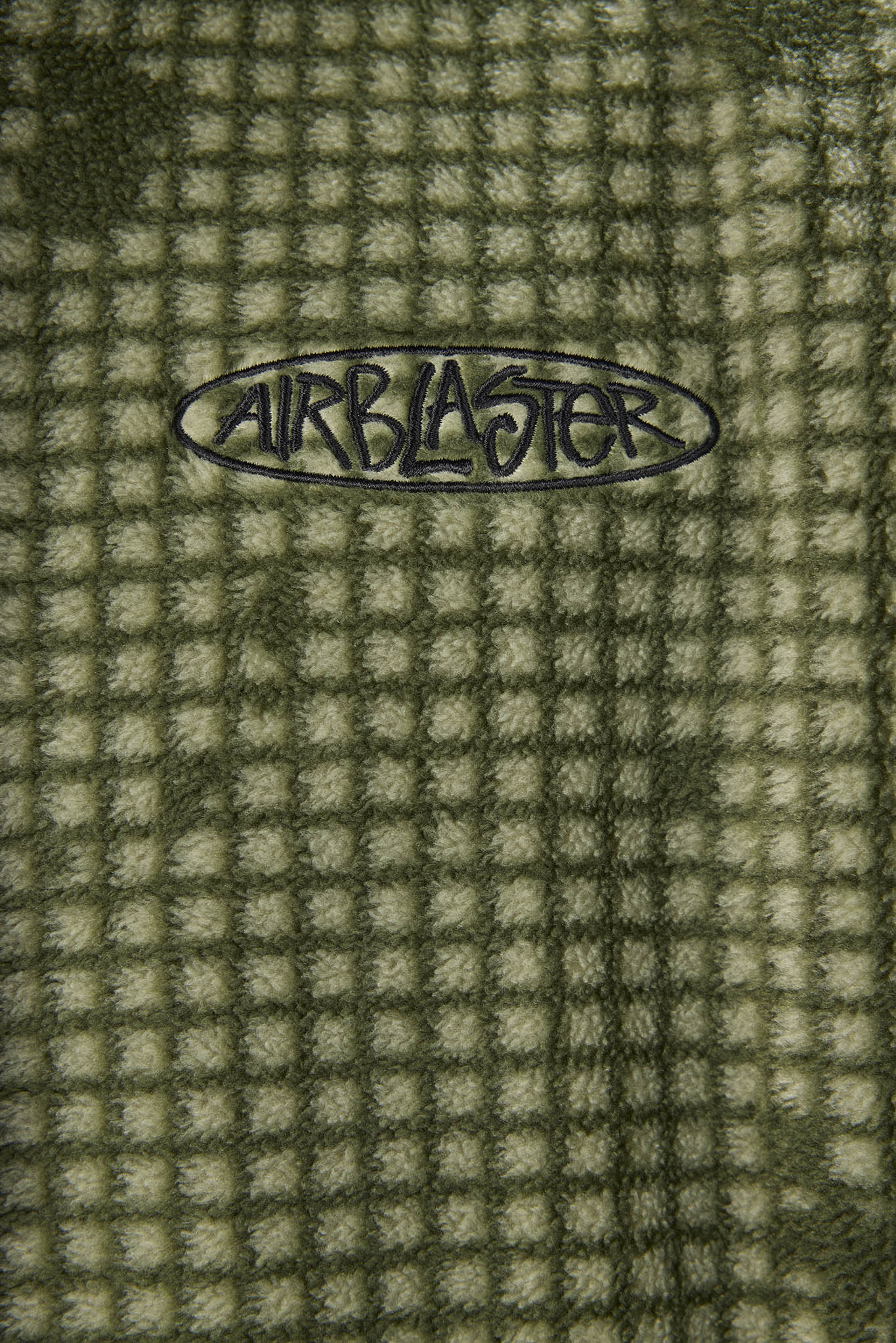 Close-up of a textured waffle fleece blanket in olive green and beige, featuring a black Airblaster logo embroidered in the center.
