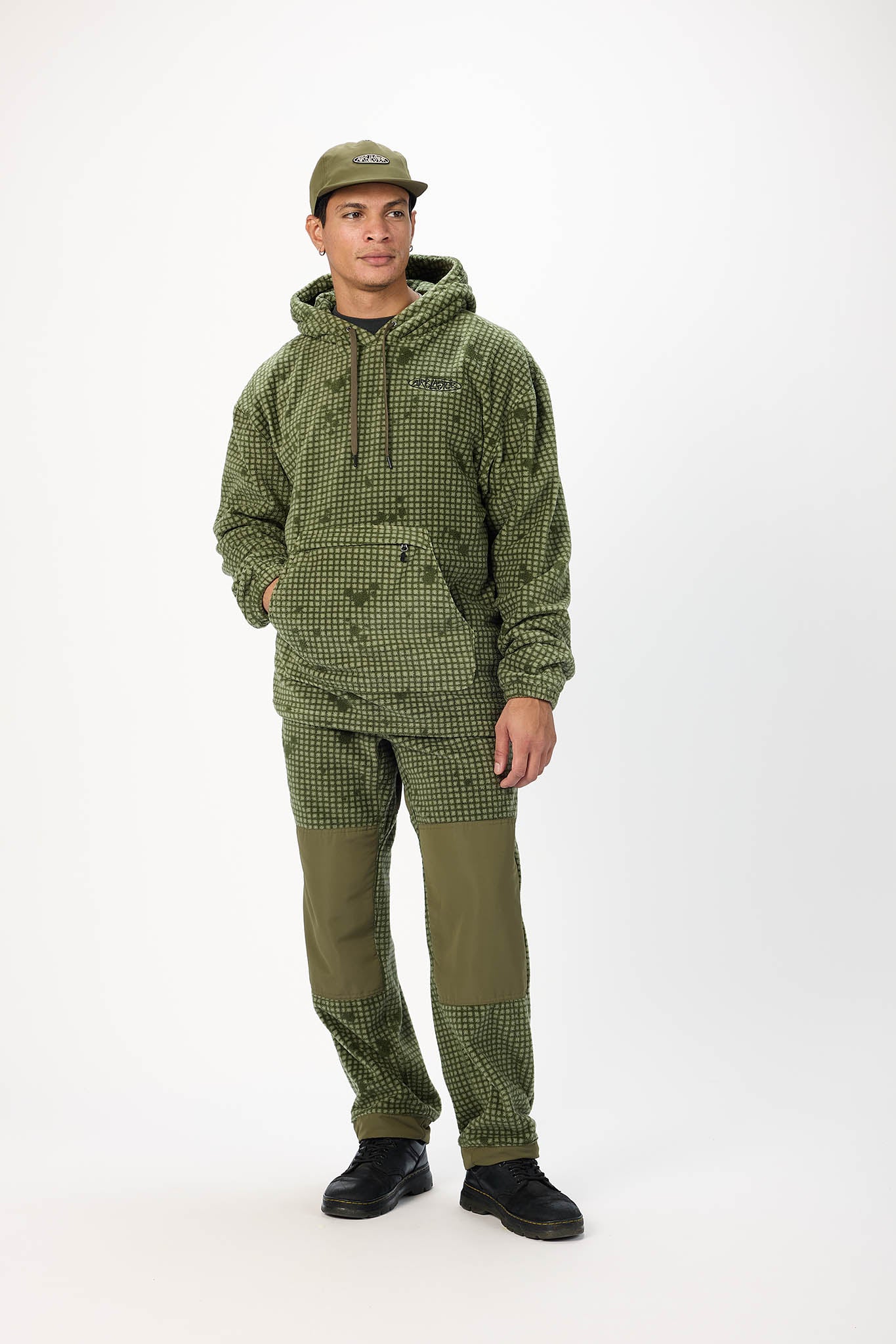 A man modeling an olive green hooded pullover jacket and matching pants with a small-scale checkered pattern and solid olive green knee patches, paired with a matching cap and black shoes, against a plain white studio background.