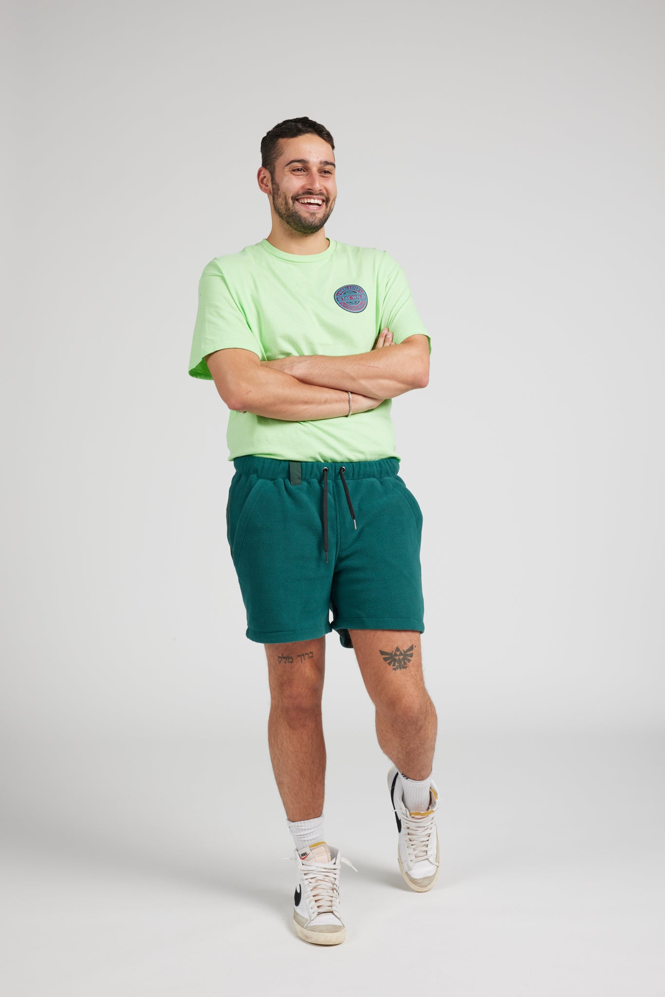 A man modeling a light green crewneck t-shirt with a circular chest graphic and dark teal drawstring shorts, paired with white socks and white sneakers, against a light gray studio background.