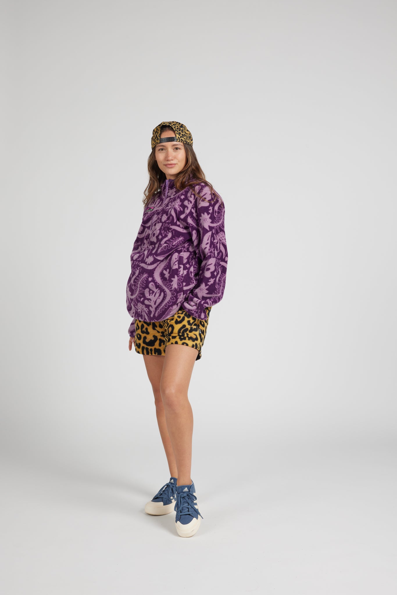 A model wearing a purple and black paisley patterned sweatshirt, yellow and black leopard print shorts, a leopard print beanie, and blue sneakers, standing against a light gray studio background.