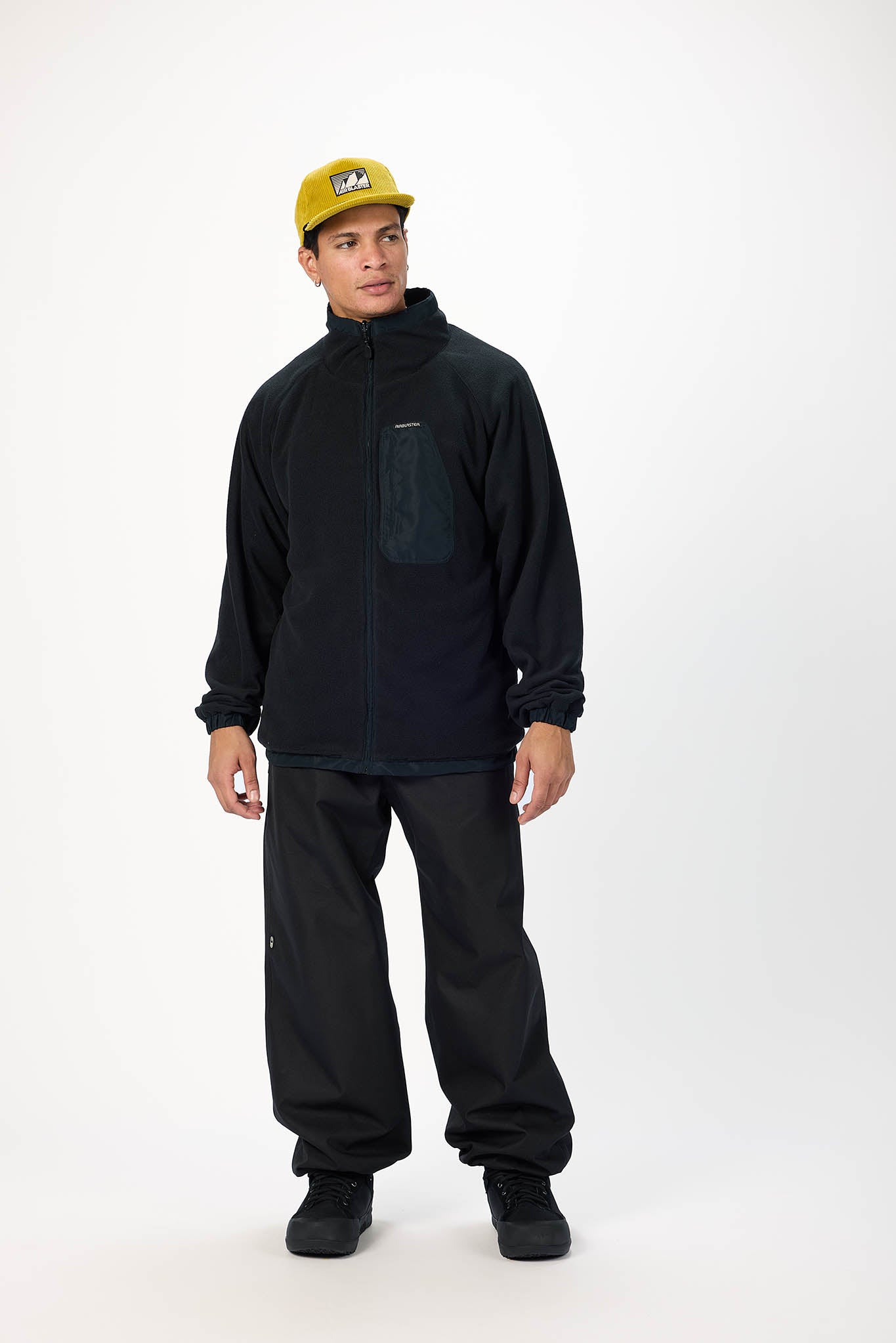 A man modeling a black fleece jacket and black pants, wearing a yellow cap, standing against a plain white studio background.
