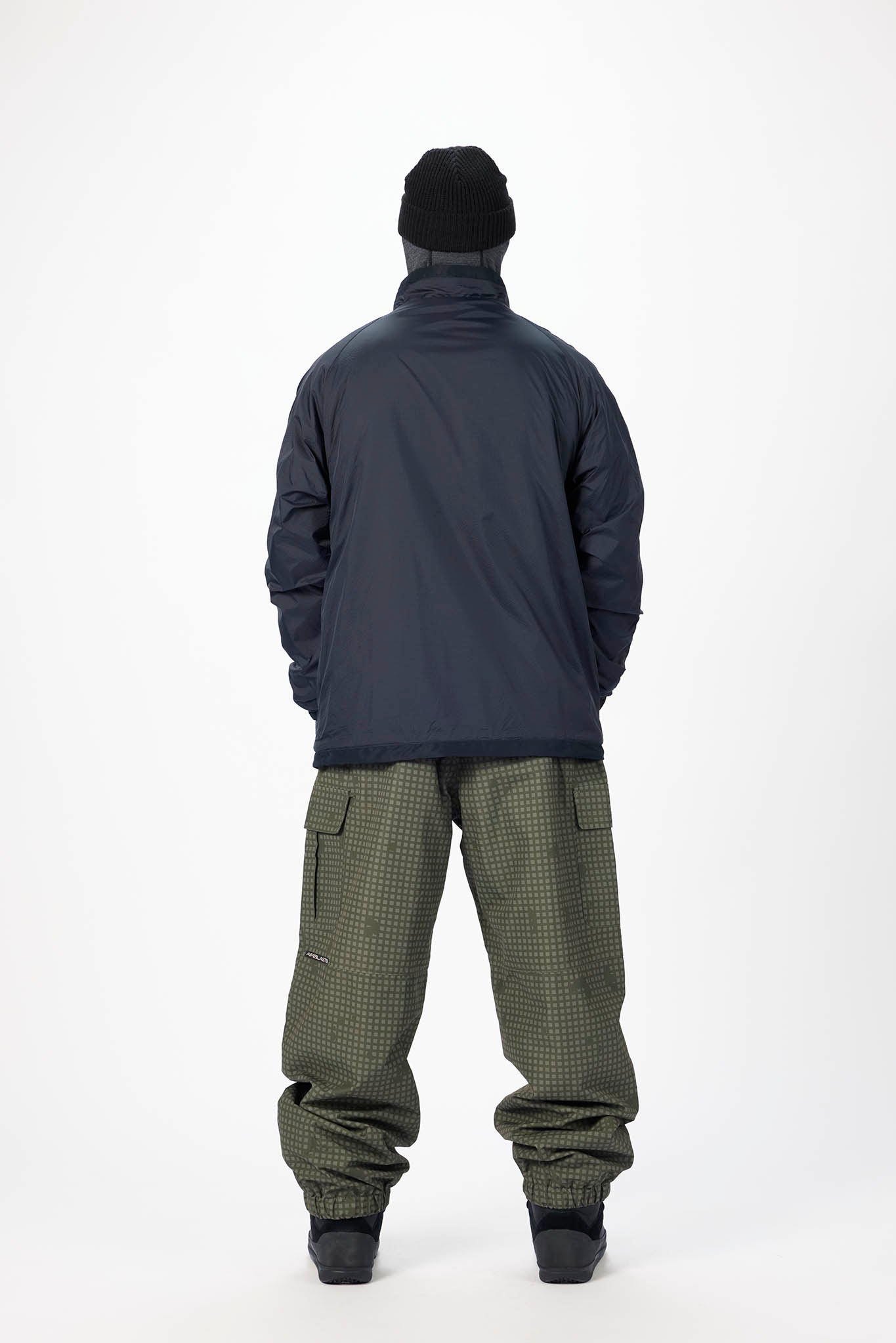 A person shown from the back wearing a dark navy jacket, olive green patterned pants, a black beanie, and black boots, standing against a plain white studio background.