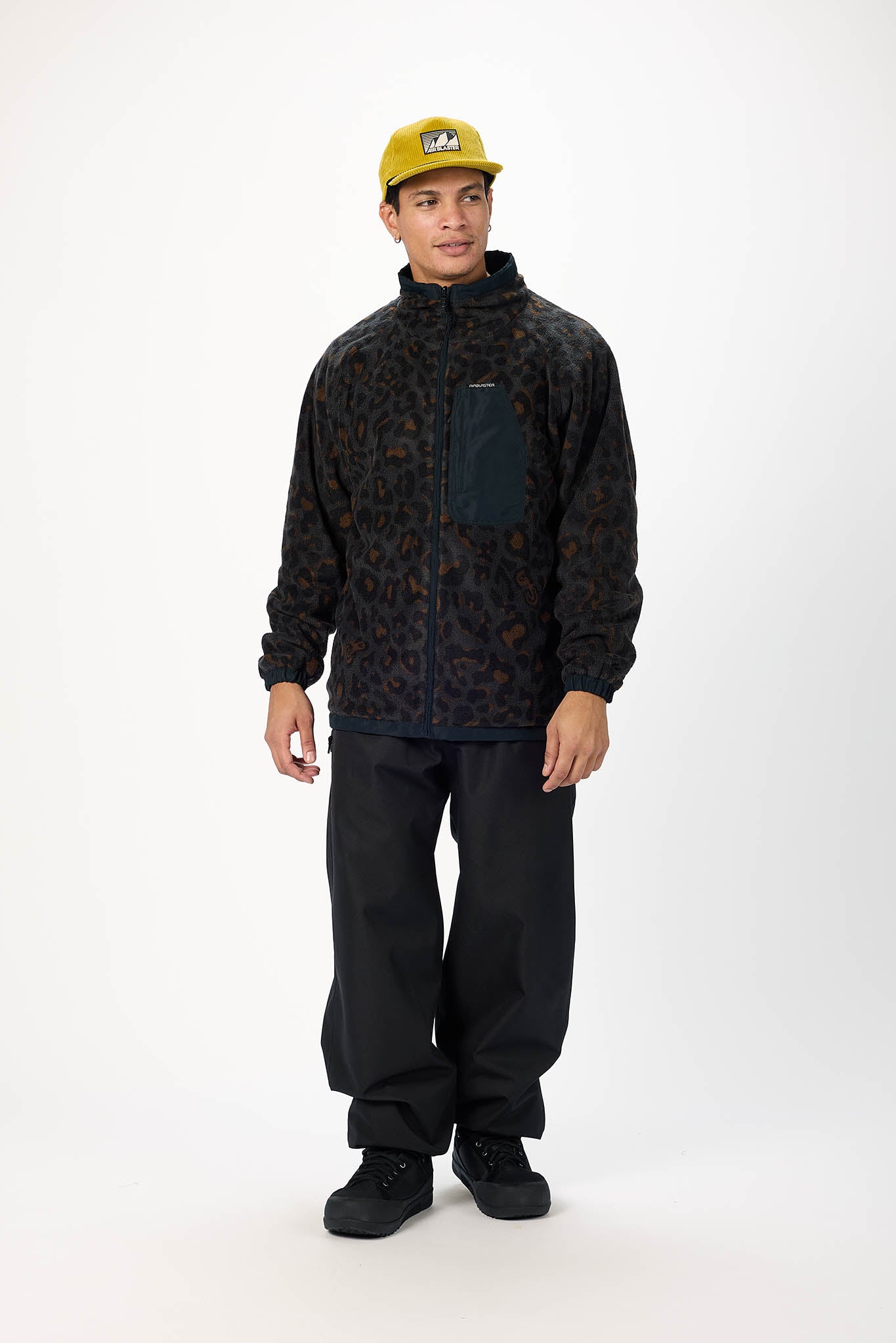 A man modeling a black and brown leopard-print fleece jacket with a dark chest pocket, paired with black pants and a yellow cap, against a plain white studio background.