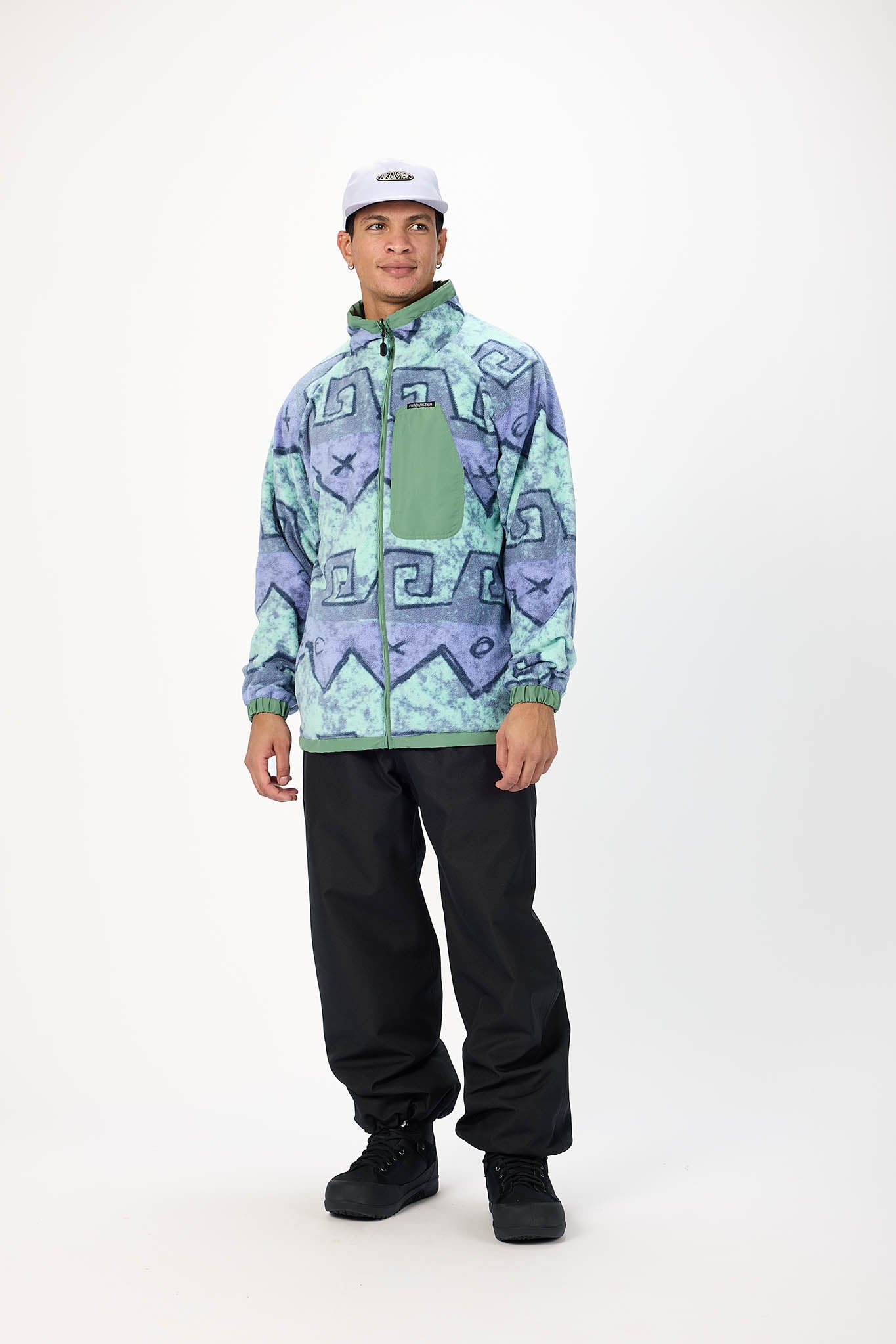 A man modeling a patterned fleece jacket with a green chest pocket, black snow pants, and a white cap, standing against a plain white studio background.